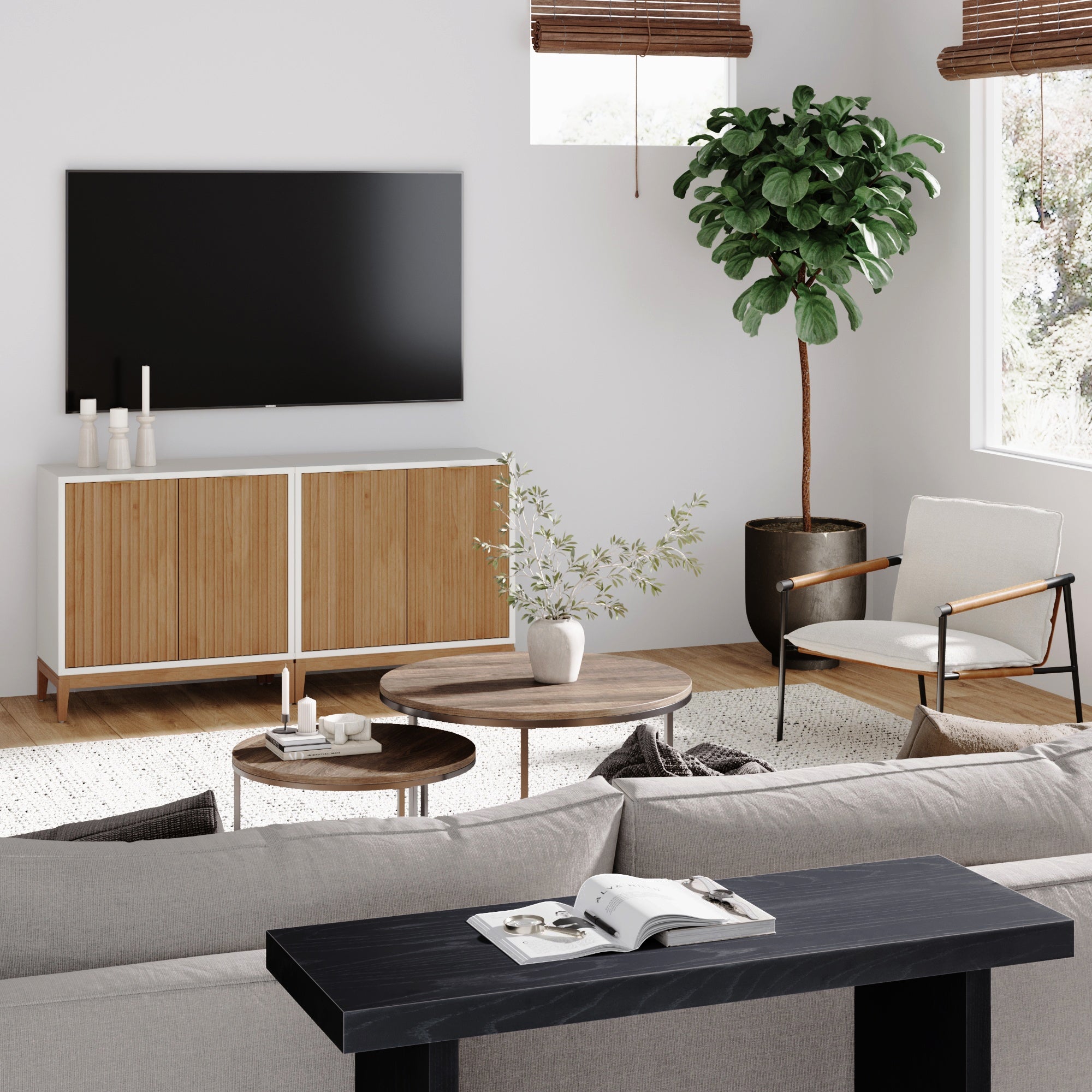 A modern living room with a wall-mounted TV, a wooden cabinet, a large potted plant, round coffee tables, an armchair, and a gray sofa. Sunlight streams through windows with bamboo shades.