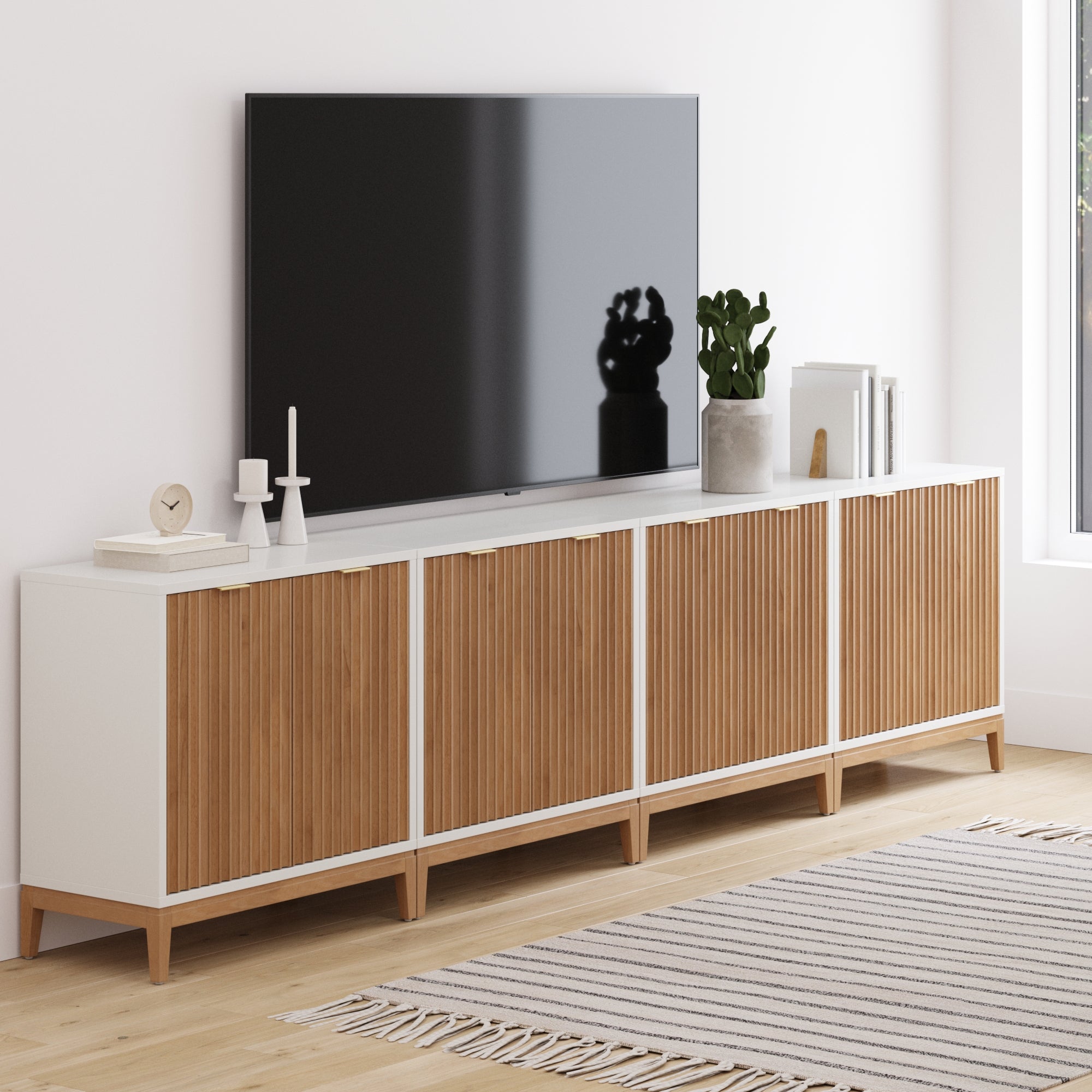 A modern living room features a large TV on the Nathan James Mid-Century Large Fluted Credenza Console Table in white, styled with candles, a clock, books, and a potted plant. A light wood floor and striped rug complete the look.