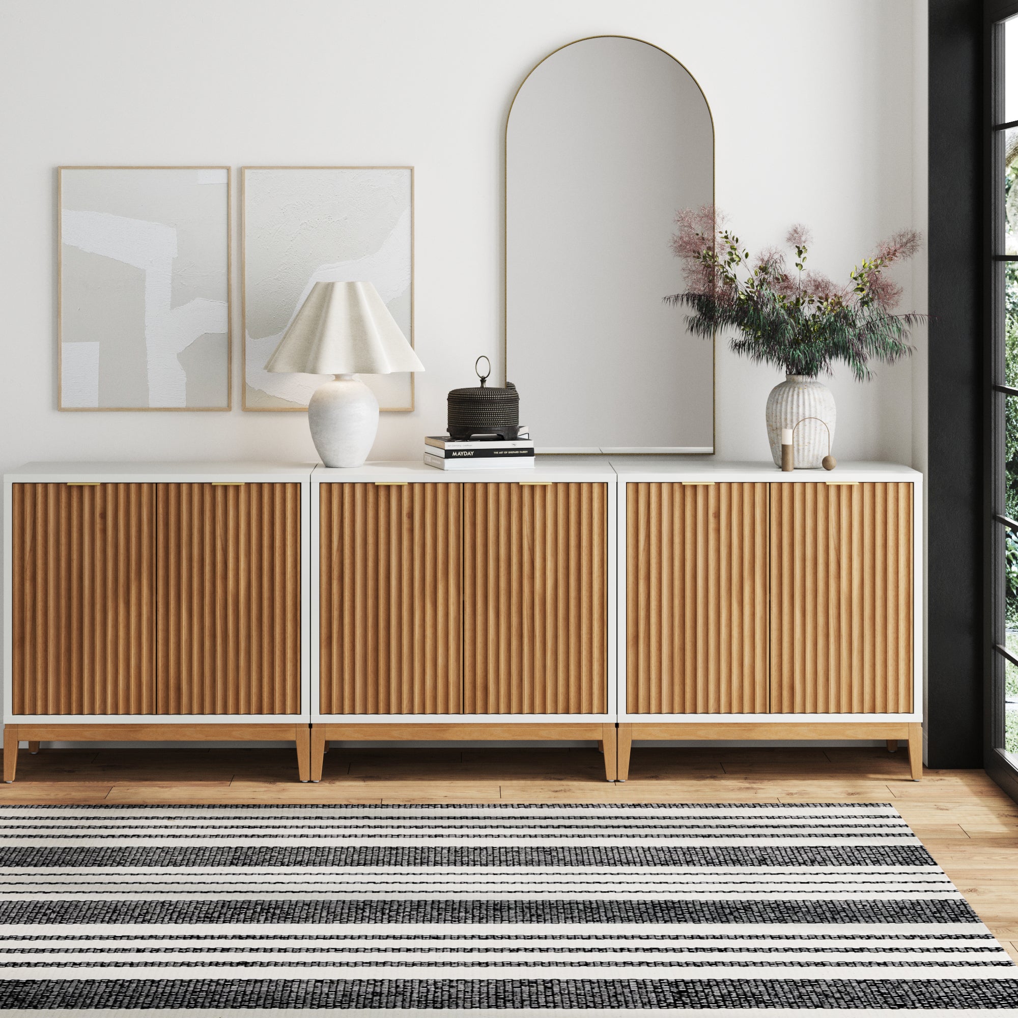 The Nathan James Mid-Century Fluted TV Console Credenza in white (Set of 3) sits against a white wall, topped with a lamp, books, a flower vase, and a round-edged mirror. Abstract art hangs above and a striped rug is on the floor.