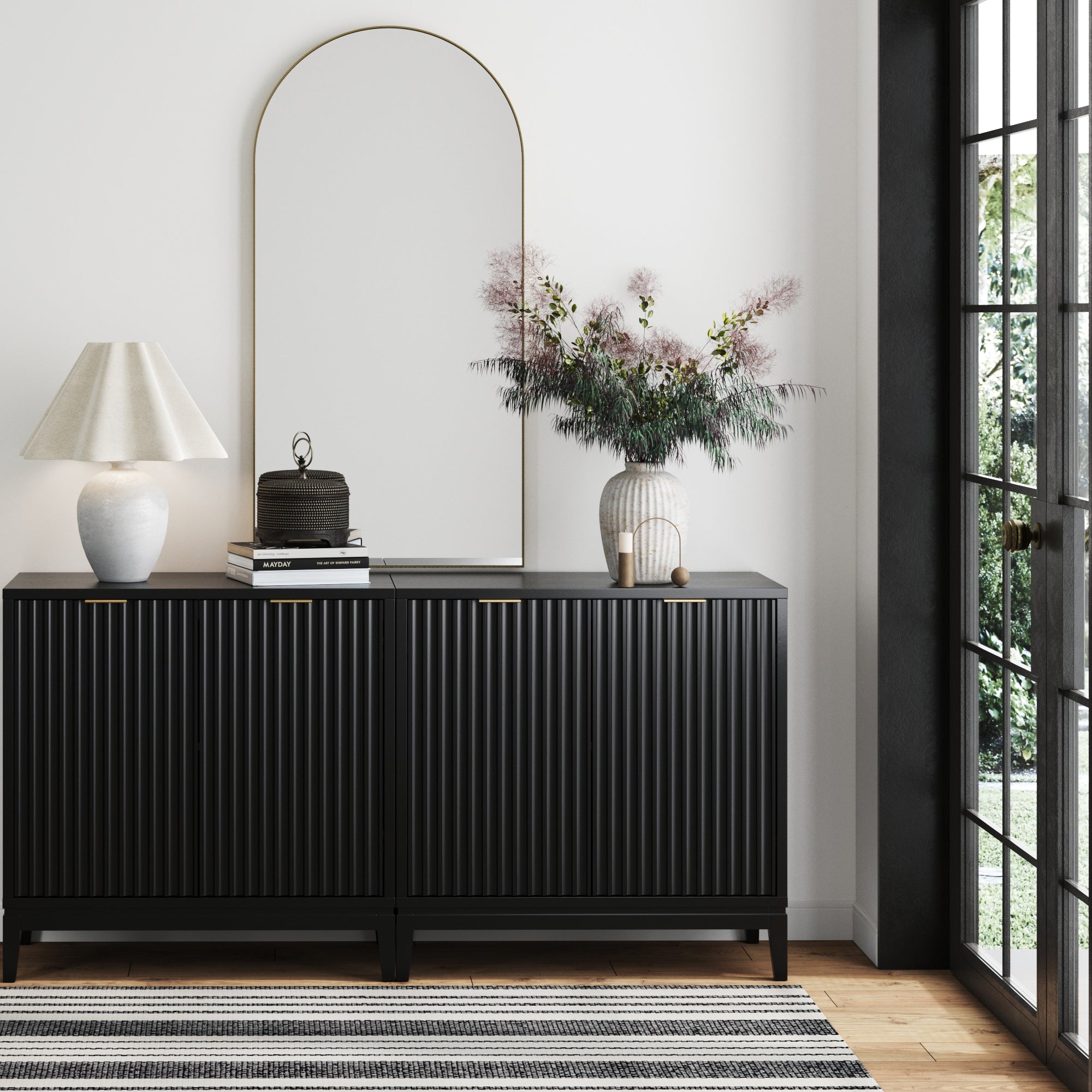 A modern interior features the Nathan James Mid-Century Fluted TV Console Credenza in black (set of 2), adorned with a white lamp, books, decorative box, and floral vase. Above hangs a large arched mirror; sunlight shines beside a striped rug.
