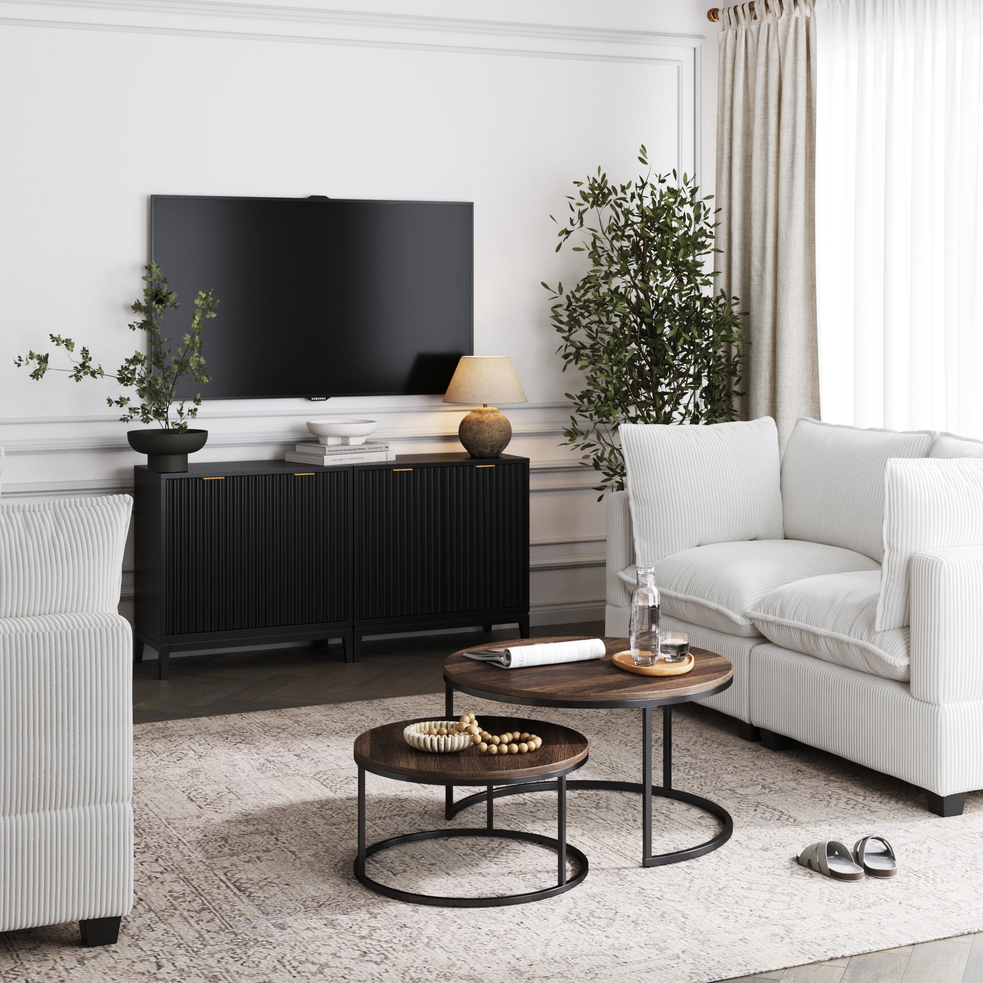 A modern living room features white sofas, a patterned rug, round wood coffee tables, a wall-mounted TV, two Nathan James Mid-Century Fluted TV Console Credenzas in black oak finish, potted plants, a lamp, and light curtains over the window.