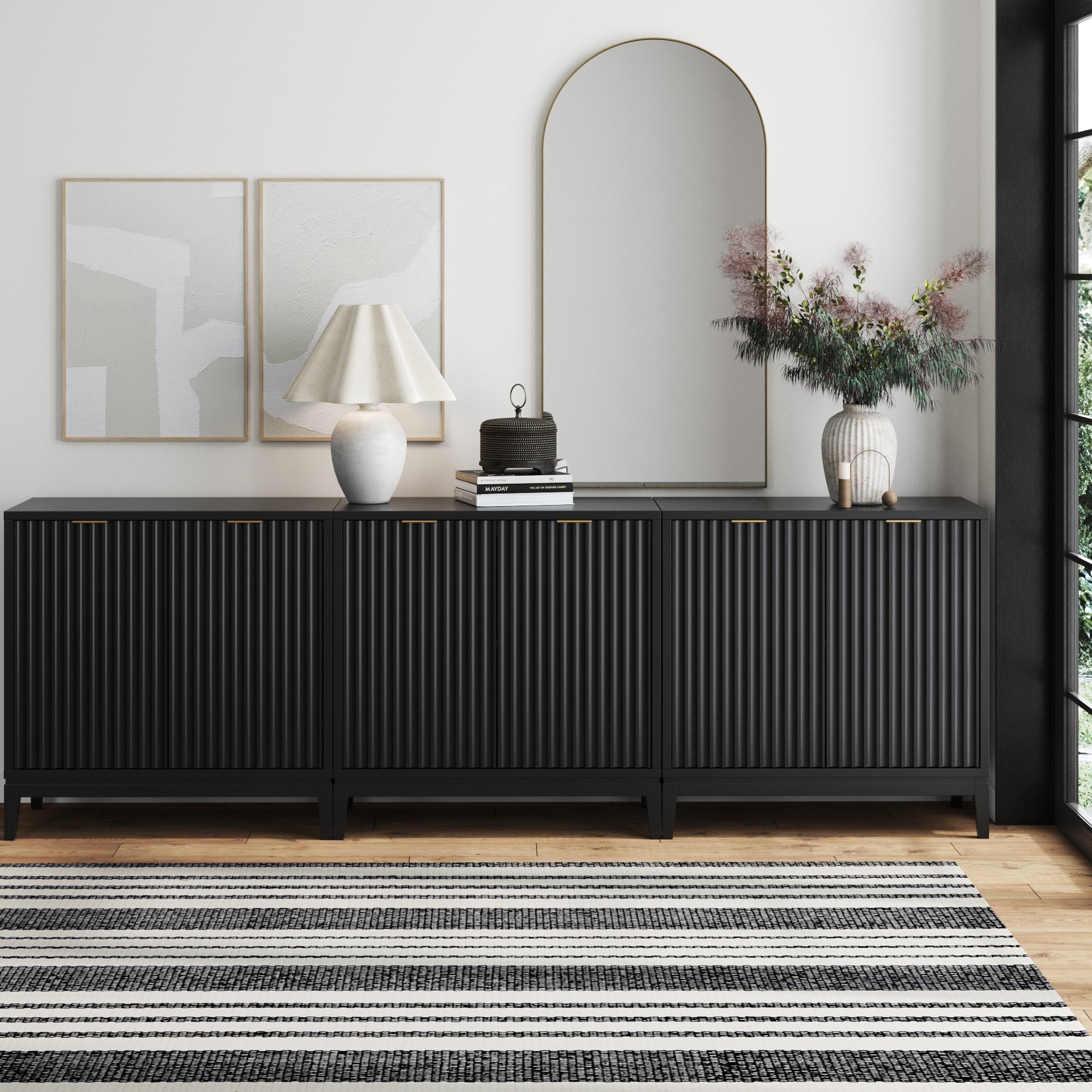 The Nathan James Mid-Century Fluted TV Console Credenza Black (Set of 3) stands against a white wall, topped with a lamp, books, flowers in a vase, and an arched mirror. Two abstract artworks are displayed beside the mirror above the credenza.