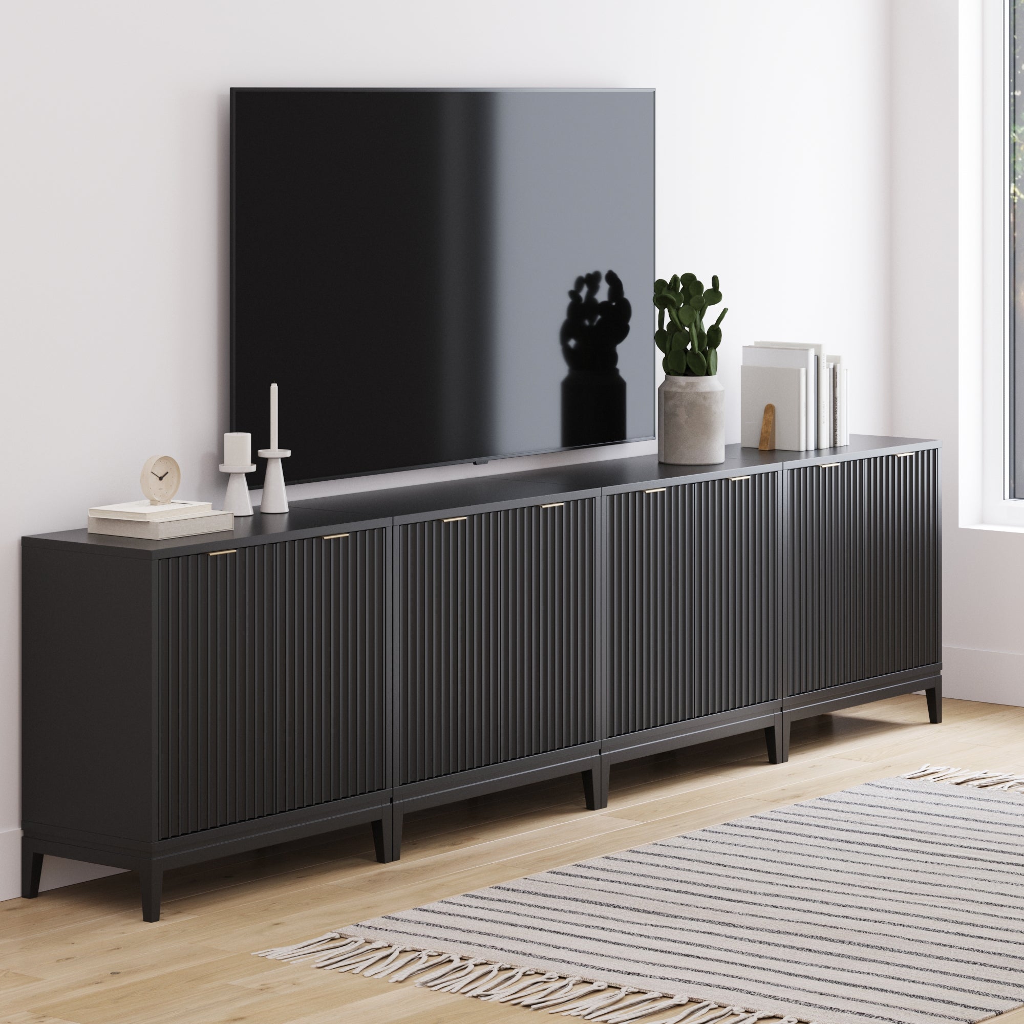 A modern living room features a large TV on the Nathan James Mid-Century Large Fluted Credenza Console Table Black (Set of 4), displaying decor, books, a plant, and candles. Sunlight shines onto a striped rug over wooden floors.