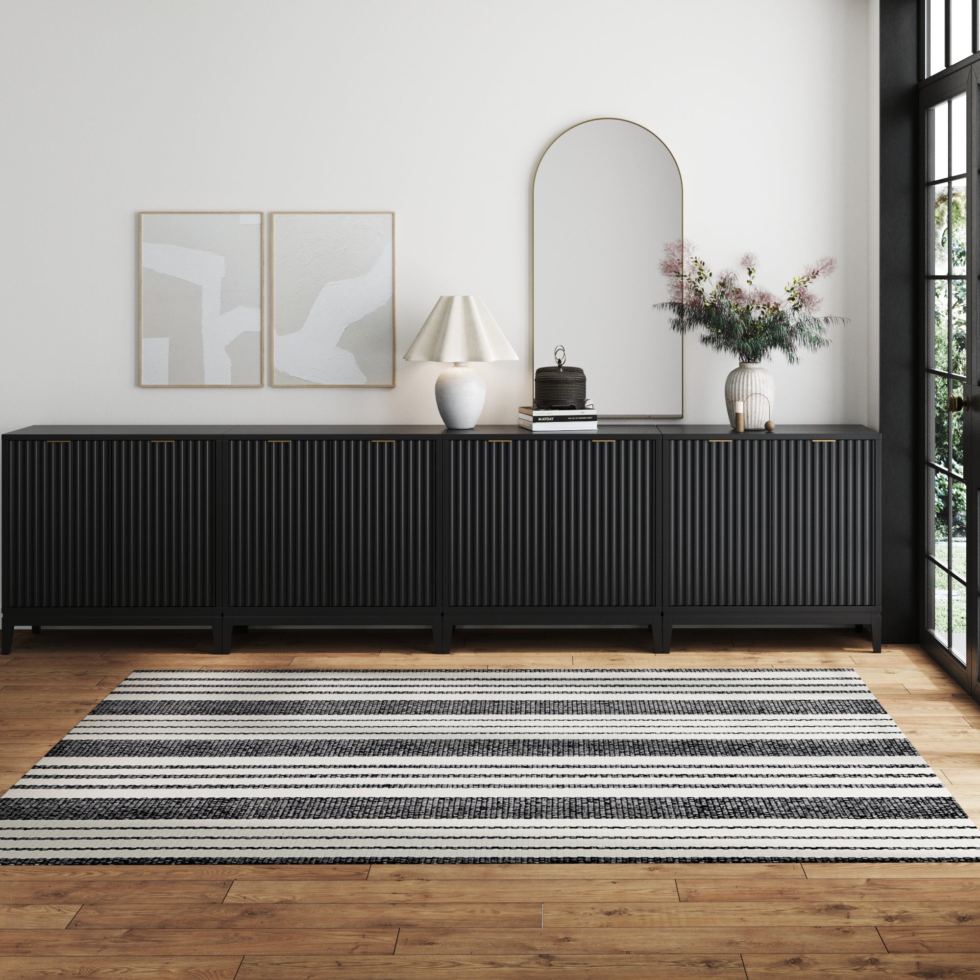 A minimalist living room with a Nathan James Mid-Century Large Fluted Credenza Console Table in black, two abstract artworks, table lamp, arched mirror, floral vase, and a black-and-white striped rug on the wooden floor.