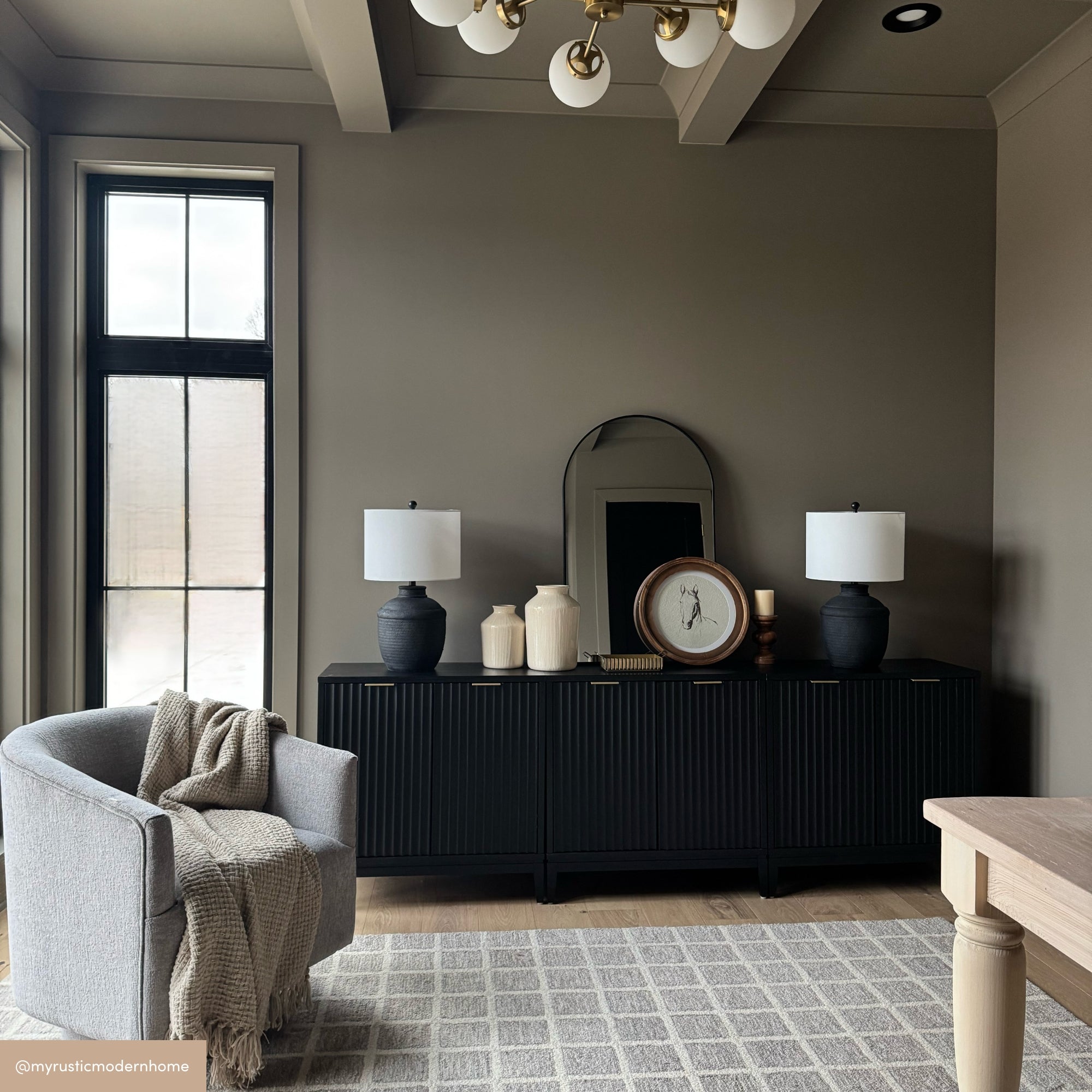 A modern living room with taupe walls, a gray upholstered chair with a beige throw, a checkered rug, and a black sideboard topped with two black lamps, a round mirror, decorative vases, and a large clock.