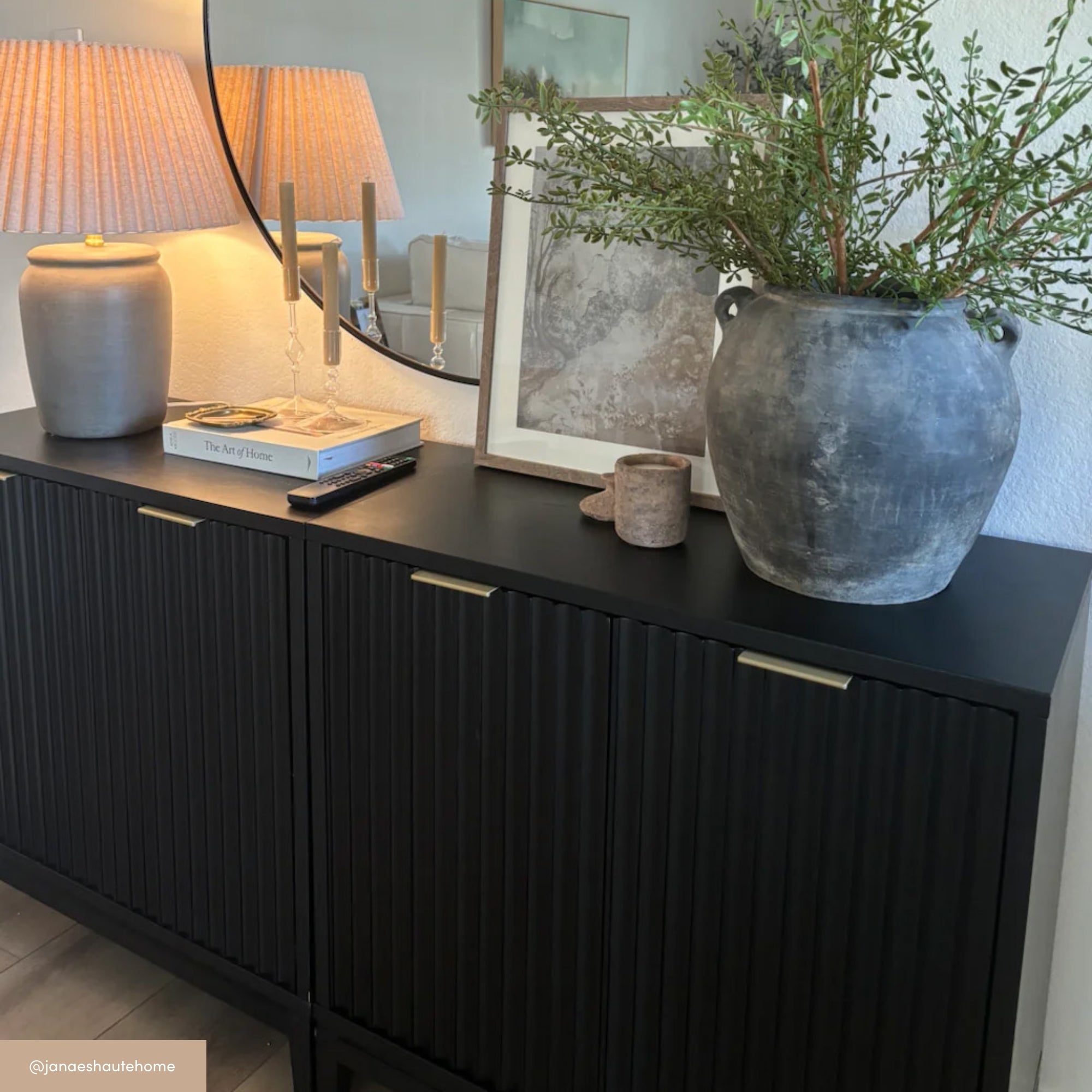 A black sideboard with vertical grooves holds a gray vase with green foliage, a small cup, a framed art print, a lamp with a beige shade, two candles, and a stack of books, positioned beneath a wall mirror.