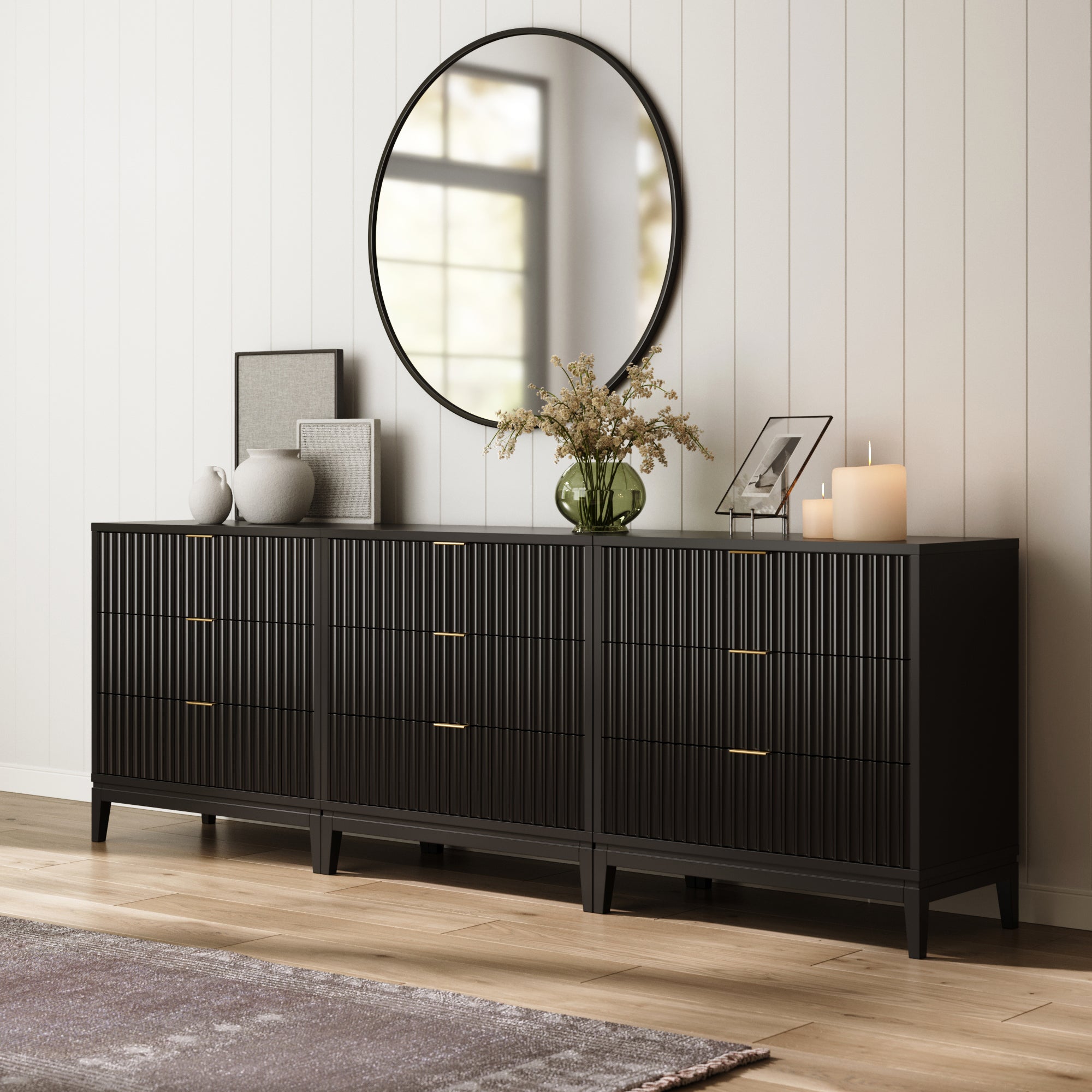 A Nathan James Mid-Century Fluted 9-Drawer Extra Large Dresser in black sits against a white paneled wall, topped with candles, a green vase of flowers, frames, and decor. A large round mirror hangs above this set of three dressers.