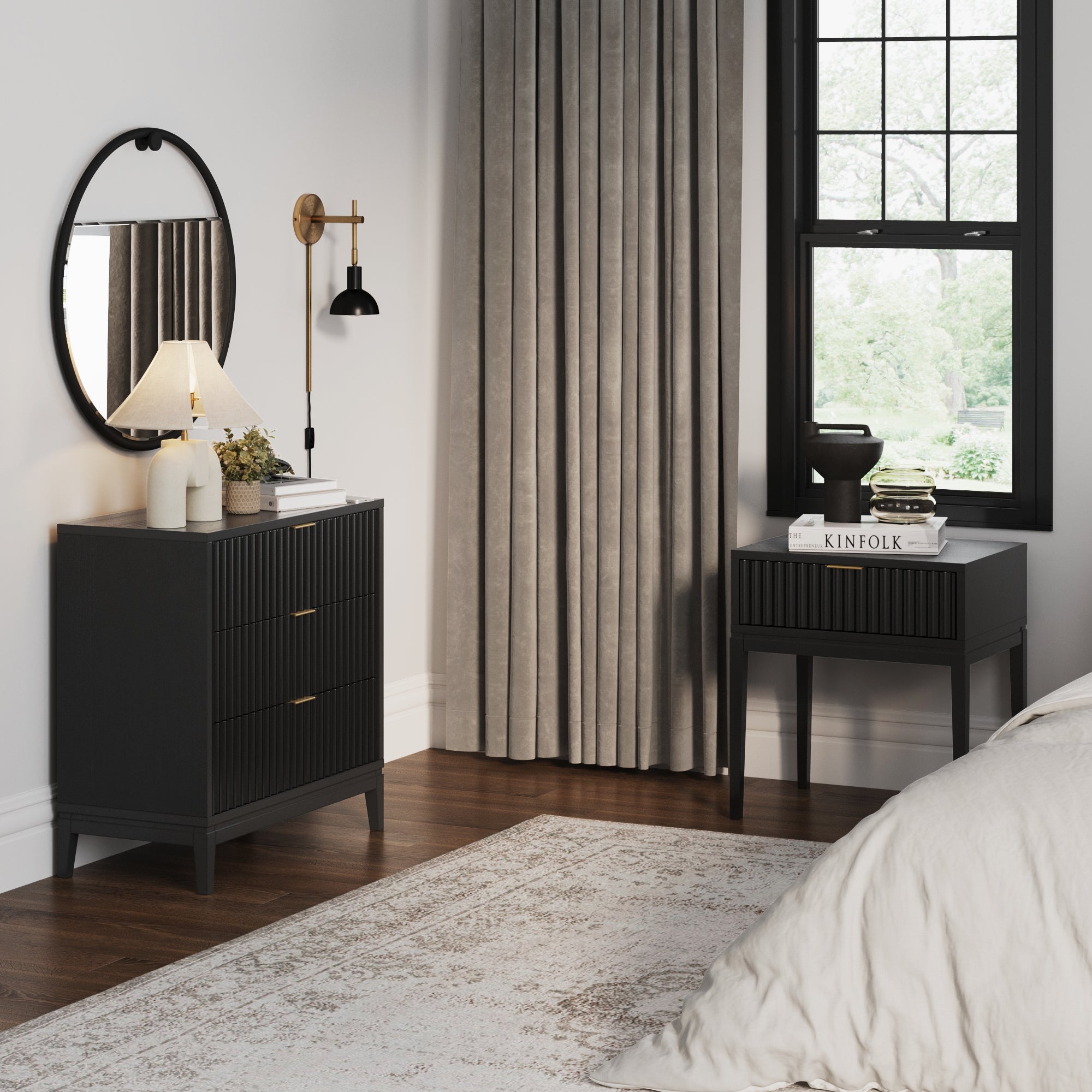 A modern bedroom features the Nathan James Mid-Century Wooden Fluted Dresser & Nightstand Set in black with soft-close drawers, styled with a round mirror, lamp, books, and decor beside white bedding and beige curtains by a large window.