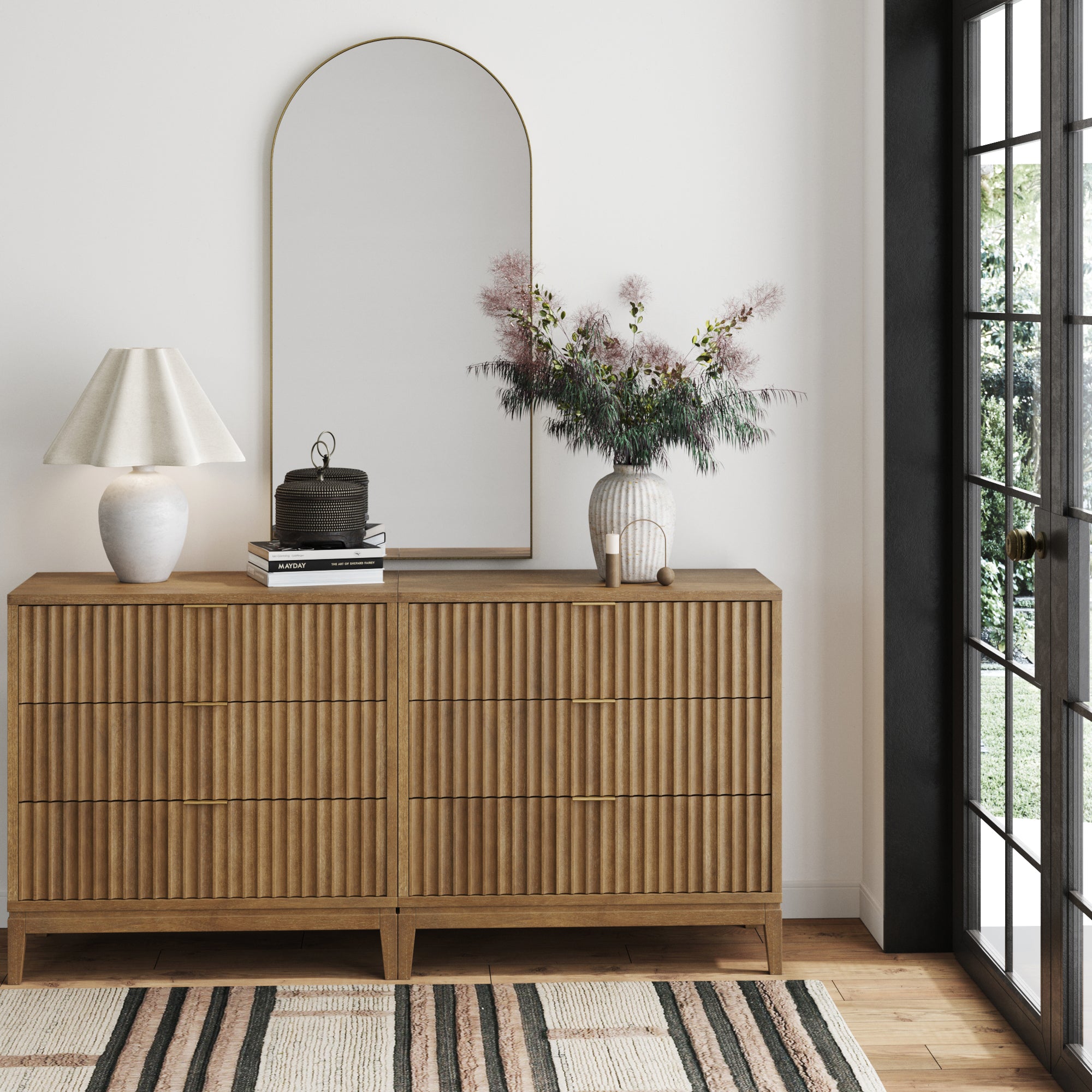 A set of two Nathan James Bohemian Solid Wood 3-Drawer Fluted Dressers in light brown, with ribbed details, stands against a white wall topped with a lamp, books, boxes, and greenery. An arched mirror hangs above near a large window.