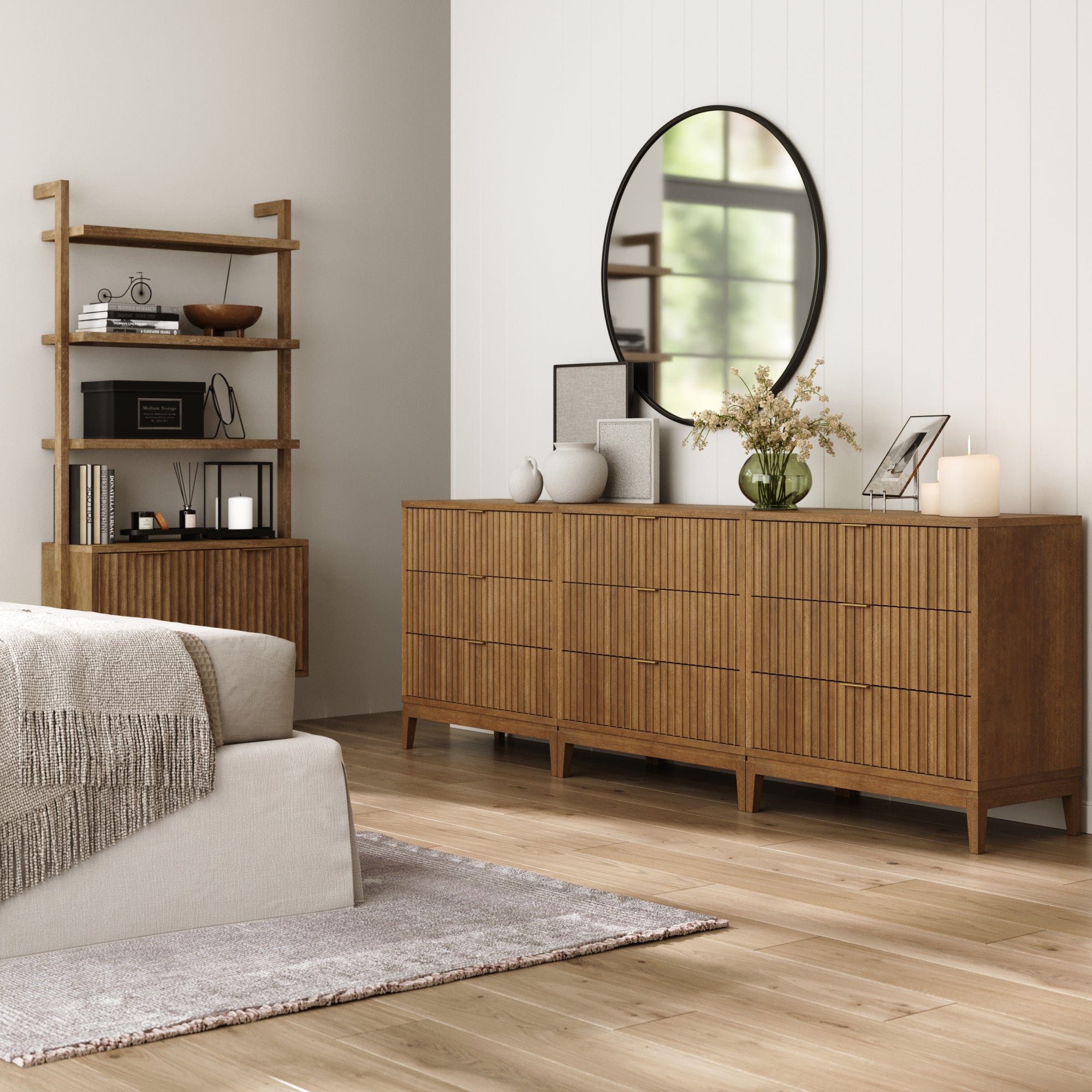 A modern bedroom with light wood floors, a beige bed and gray blanket, a Nathan James Bohemian Solid Wood 3-Drawer Fluted Dresser (Set of 3) in light brown with decor, a round mirror above, and a wooden shelf in the corner.