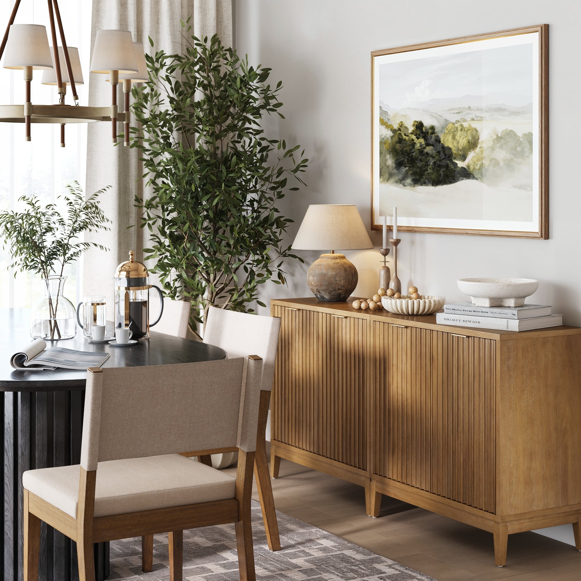A cozy dining area features light wood furniture, a round black coffee table, beige upholstered chairs, and two Nathan James Mid-Century Fluted TV Console Credenzas in light brown. Sunlight filters onto landscape art and a potted plant.