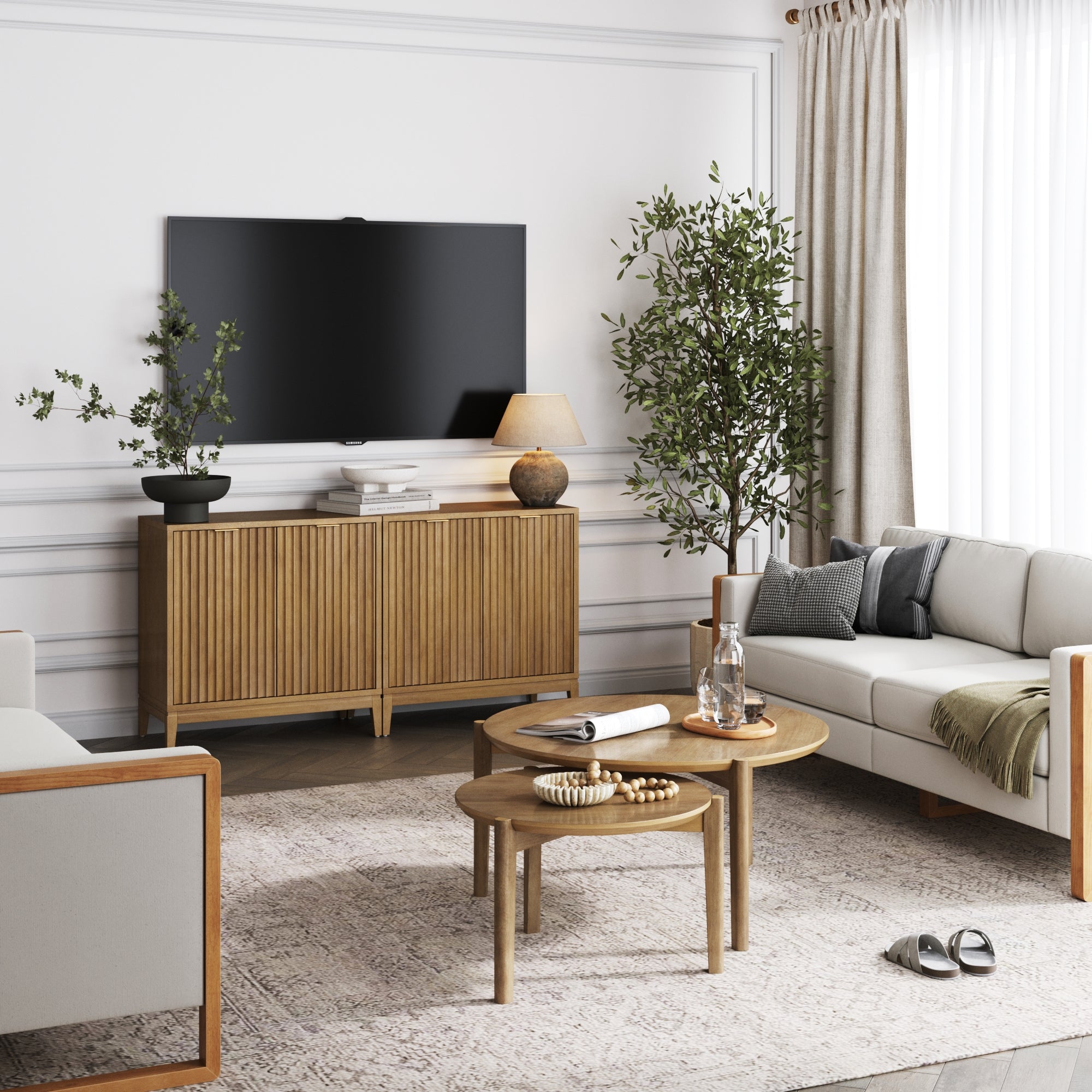 A modern living room with a wall-mounted TV, wooden sideboard, two potted plants, round nesting coffee tables, sofas, light curtains, and a patterned rug on a light floor.