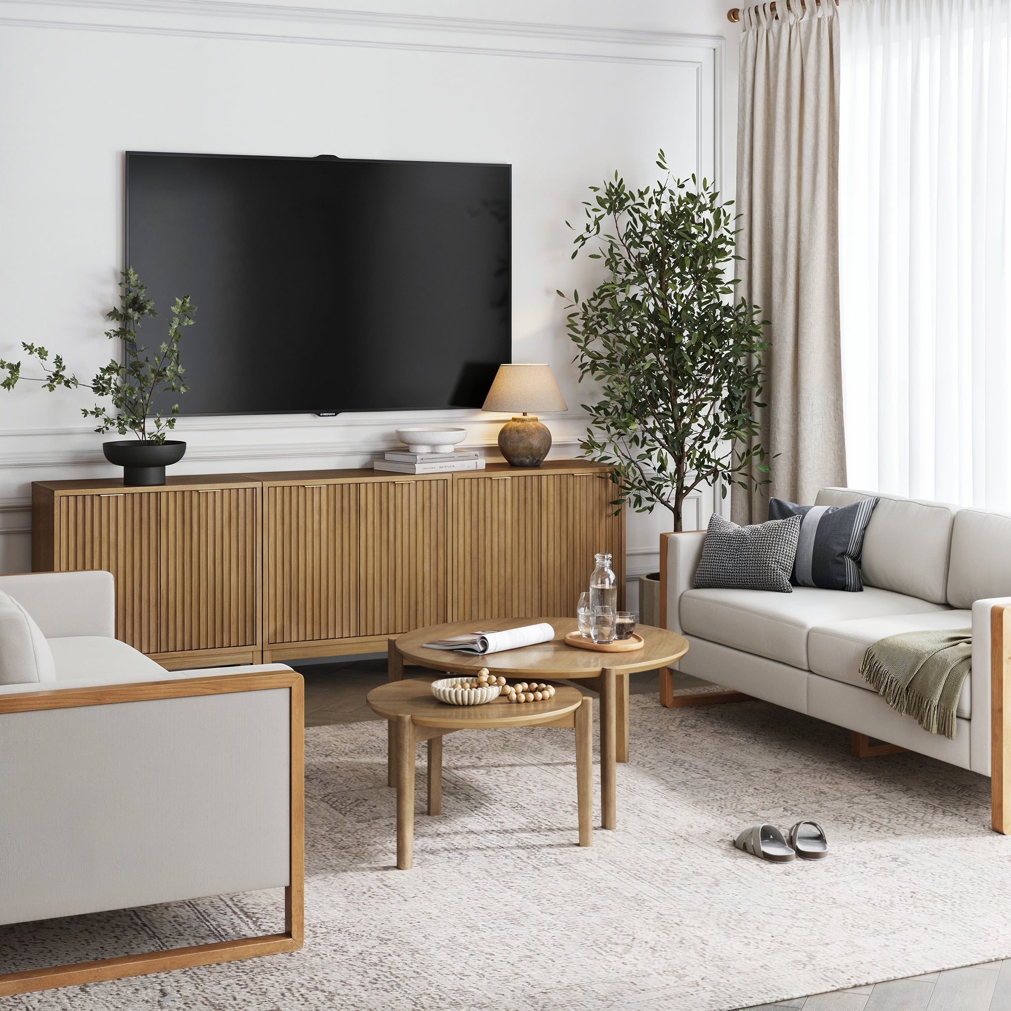 A modern living room with neutral tones features a large TV, two beige sofas, a wooden armchair, round coffee tables, potted plants, and the Nathan James Mid-Century Fluted TV Console Credenza Light Brown (Set of 3), all bathed in natural light from tall curtains.