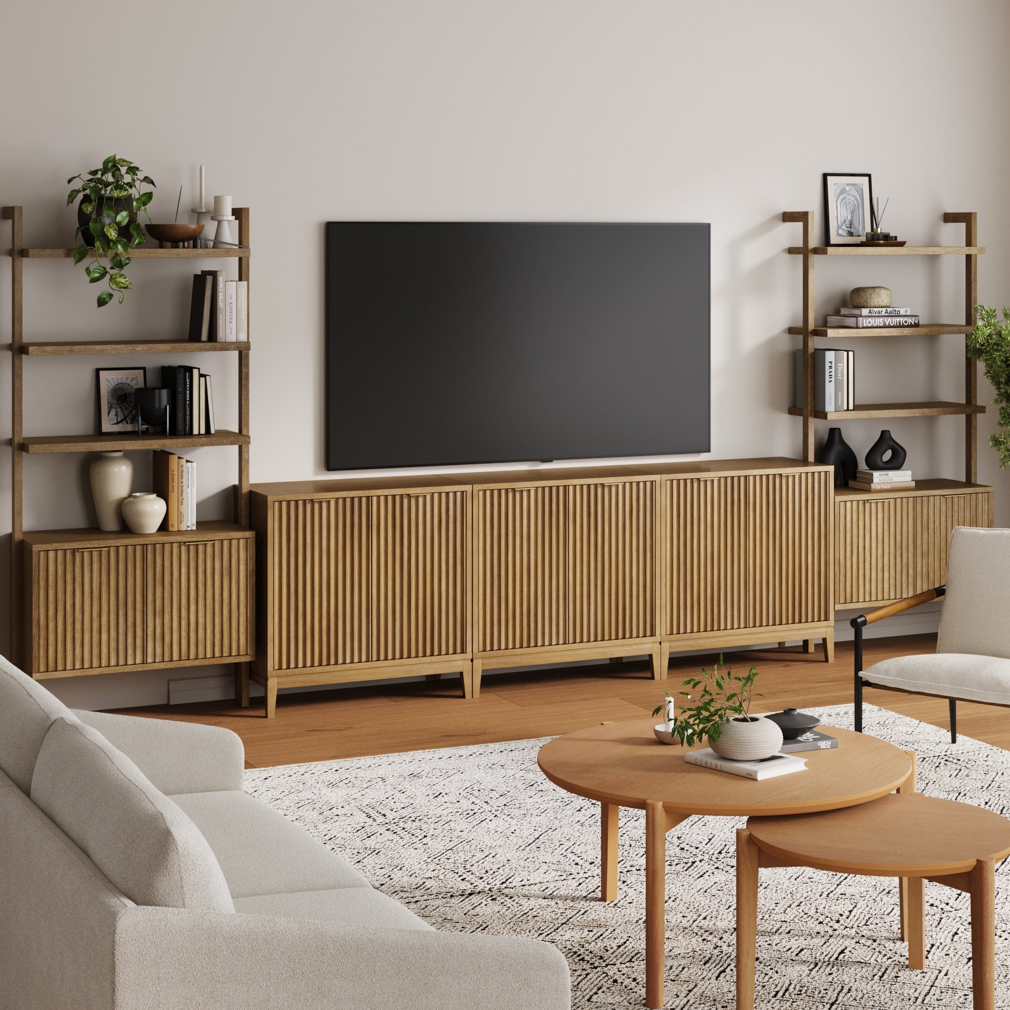 Modern living room with a Nathan James Mid-Century Entertainment Center in light brown (3 cabinets), fluted door details, large TV, shelves for plants and decor. Two light sofas and a round coffee table sit on a patterned rug.