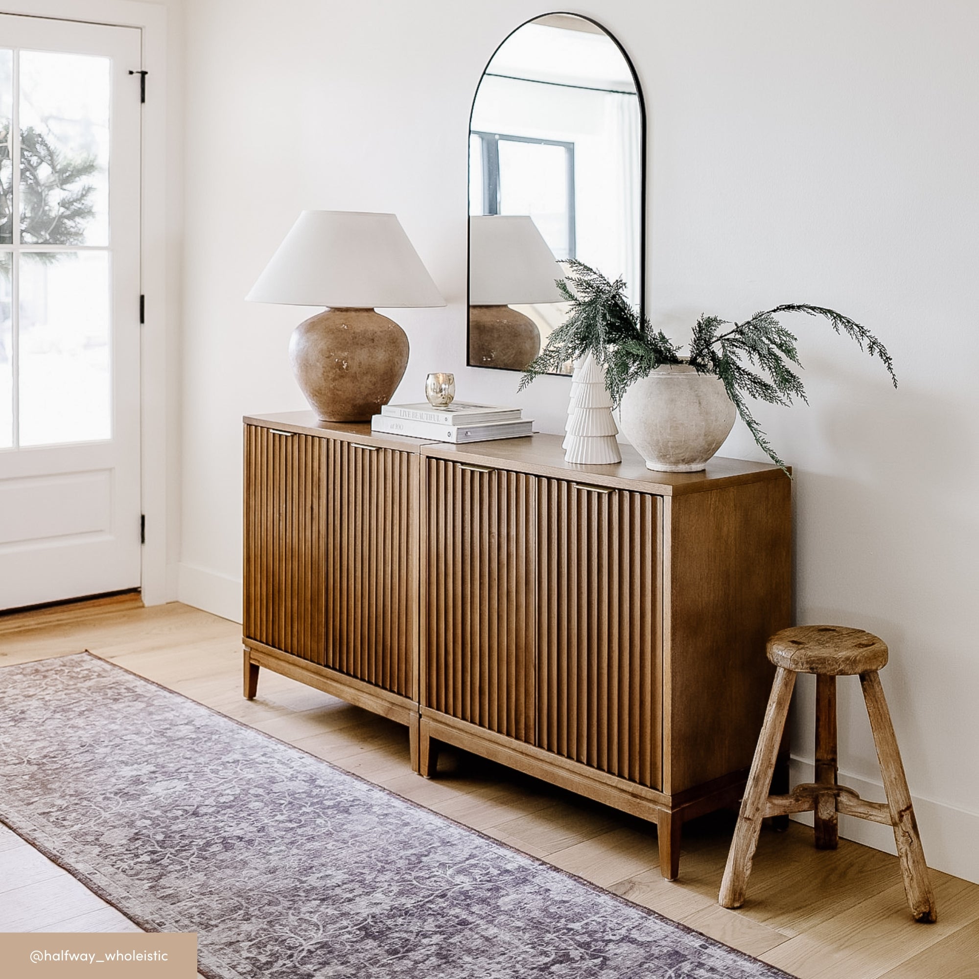 A wooden sideboard with a ribbed design stands against a white wall, topped with a tall oval mirror, books, a ceramic lamp, decorative vases, and a leafy branch. A rustic stool and a patterned rug complete the cozy entryway scene.