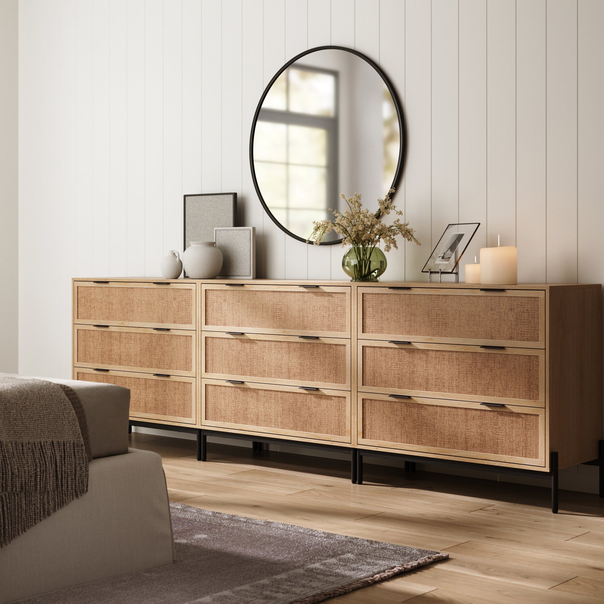A modern bedroom showcases Nathan James Rattan & Wood Dressers Light Oak (Set of 3), topped with a round mirror, vases, framed art, candles, and a plant—all against white paneled walls and wood flooring.