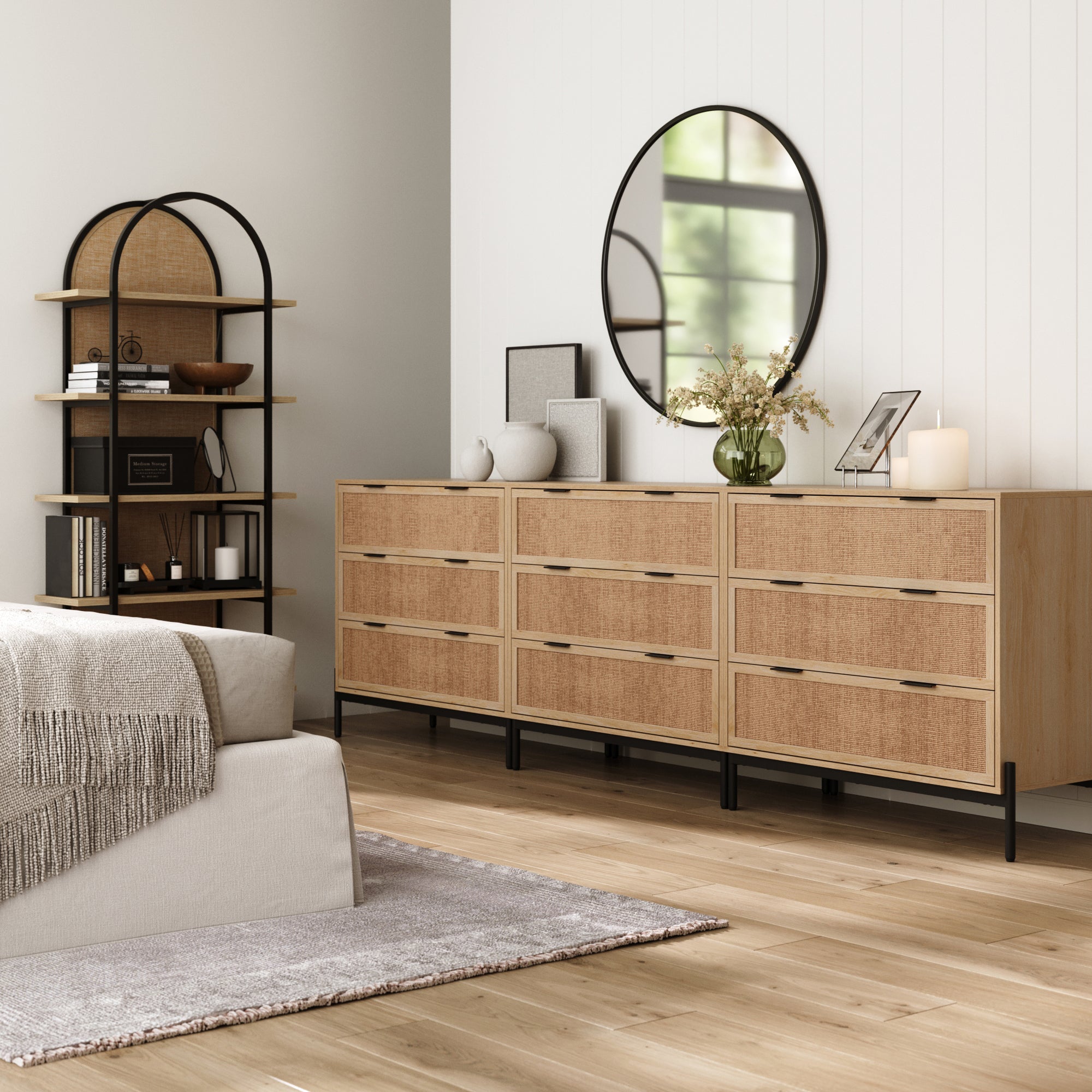 A modern bedroom has a light wood floor, beige bed with gray blanket, and Nathan James Rattan & Wood Dressers in Light Oak (Set of 3), topped with decor and a round mirror. A tall black shelving unit holds books and baskets.