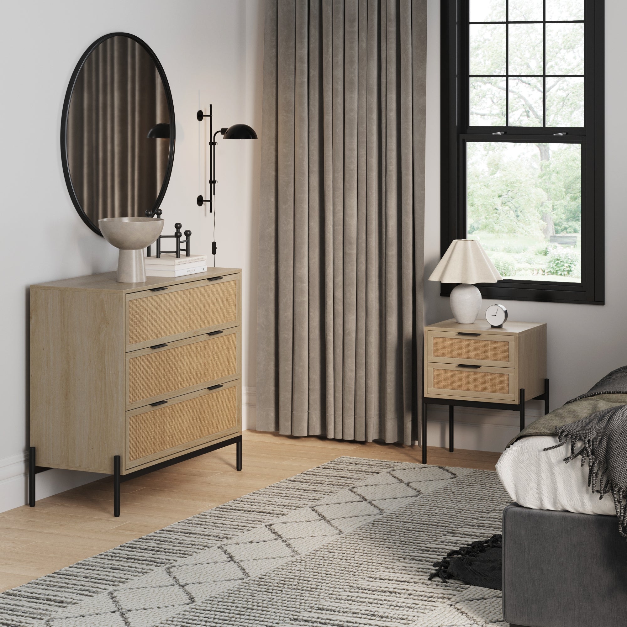A modern bedroom features Nathan James’s Oak Rattan Nightstand and Dresser Set with Drawers, a round mirror, standing lamp, beige curtains, black-framed window, bed, and a geometric-patterned rug on light wood floors.