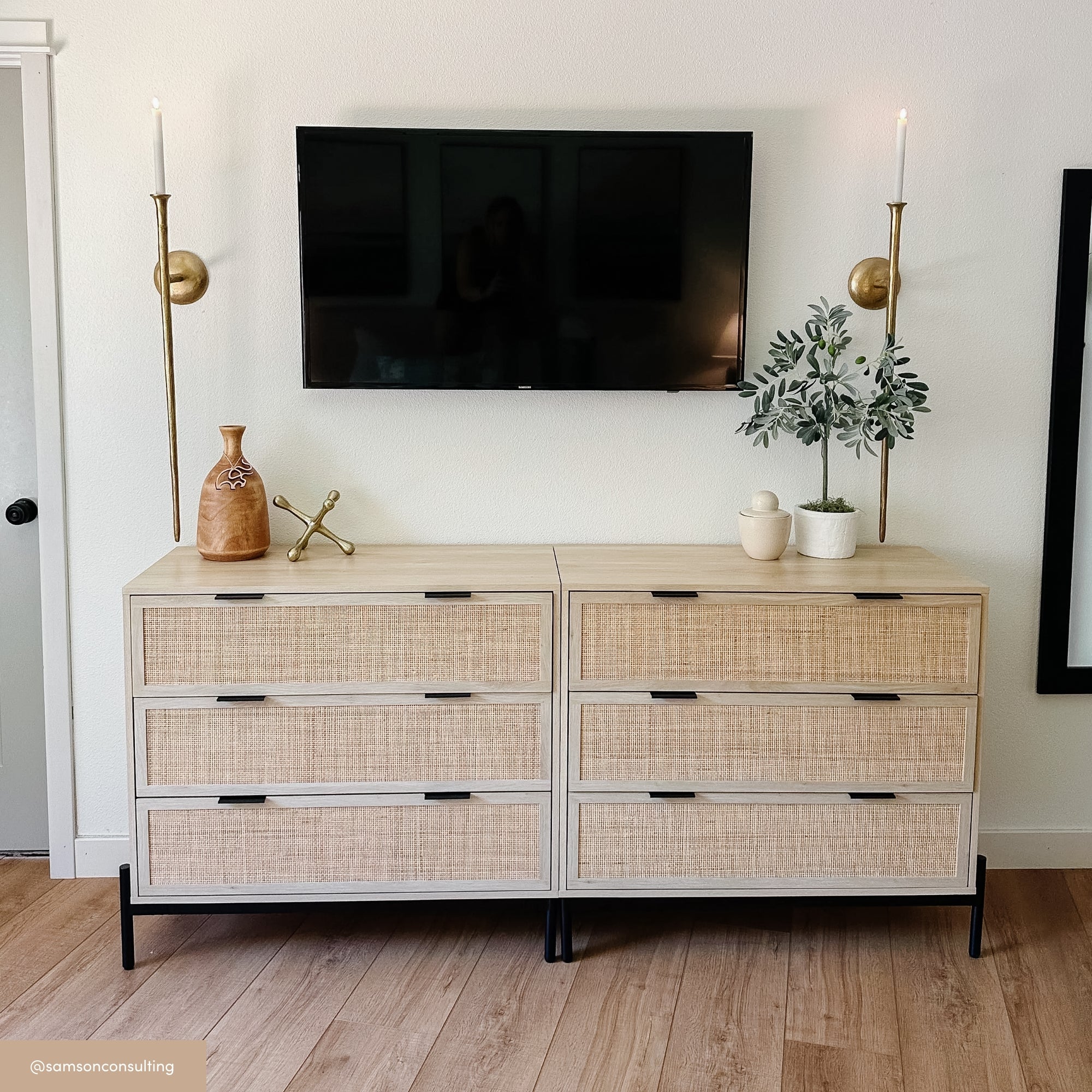 A light wood dresser with six drawers and black handles sits against a white wall. Above are wall-mounted sconces and a flatscreen TV. On the dresser are decorative items, including plants, pottery, and a gold ornament.