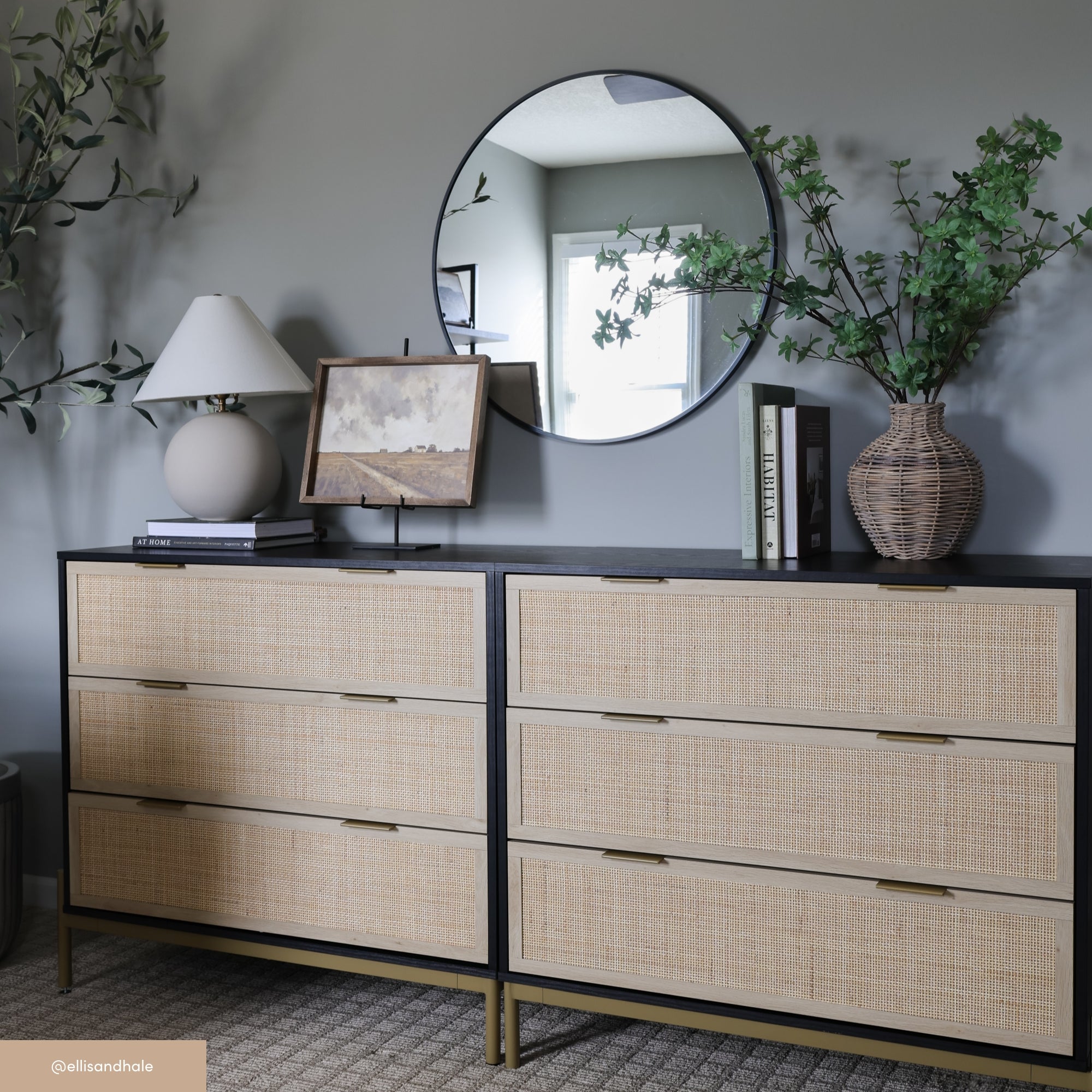 A modern dresser with six drawers features a rattan front. On top sit a round mirror, a vase with greenery, books, framed art, a ceramic lamp, and a decorative branch, all against a gray wall and carpeted floor.