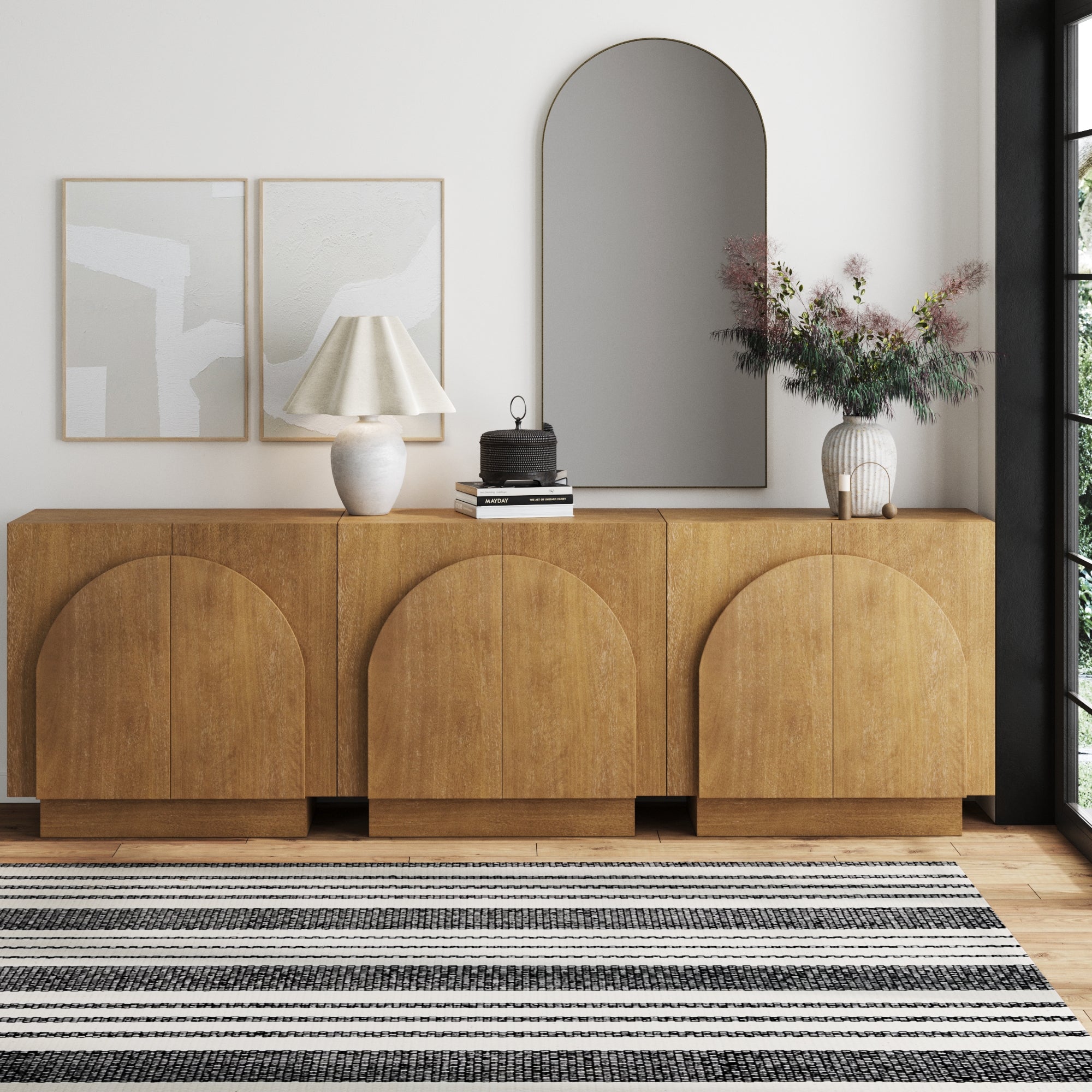 A modern living room with a Nathan James Arched TV Console Credenza Light Brown (Set of 3), adjustable shelves, abstract art, a lamp, decor accents, a vase with flowers, large arched mirror, and black and white striped rug on wood floors.
