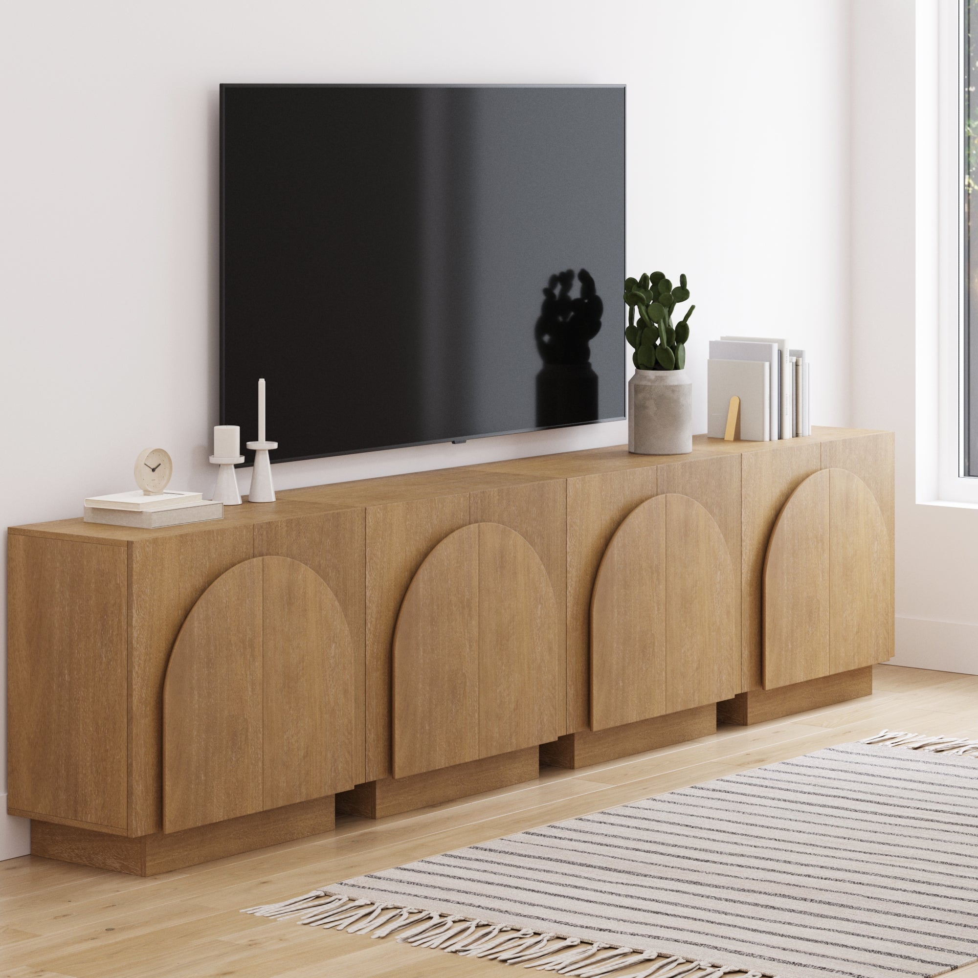 The Nathan James Large Arched TV Console Credenza in light brown (set of 4) sits beneath a wall-mounted TV, topped with candles, a clock, books, and a potted plant. A striped rug is placed on the light wooden floor nearby.