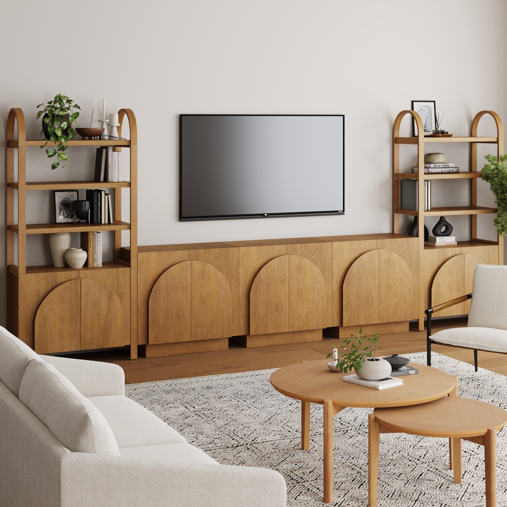 A modern living room with the Nathan James Mid-Century Modern Arched Entertainment Center, a wall-mounted TV, two tall shelves with plants and books, a round coffee table, white sofa, patterned rug, all set on wooden flooring.
