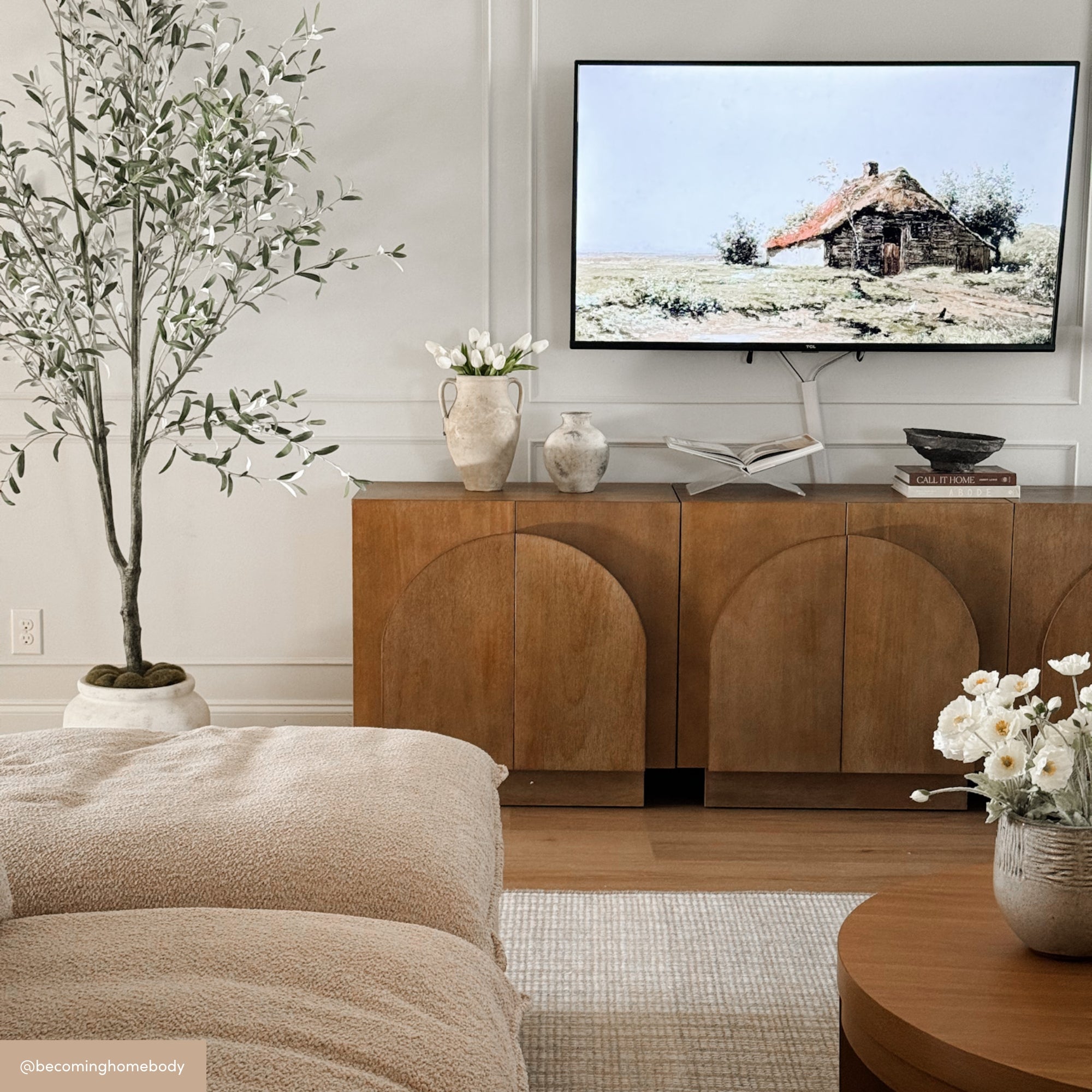 A cozy, neutral-toned living room with a large indoor plant, a soft beige sofa, wooden cabinet, coffee table with white flowers, and a TV displaying a rustic cabin scene. Decorative vases and books sit on the cabinet.