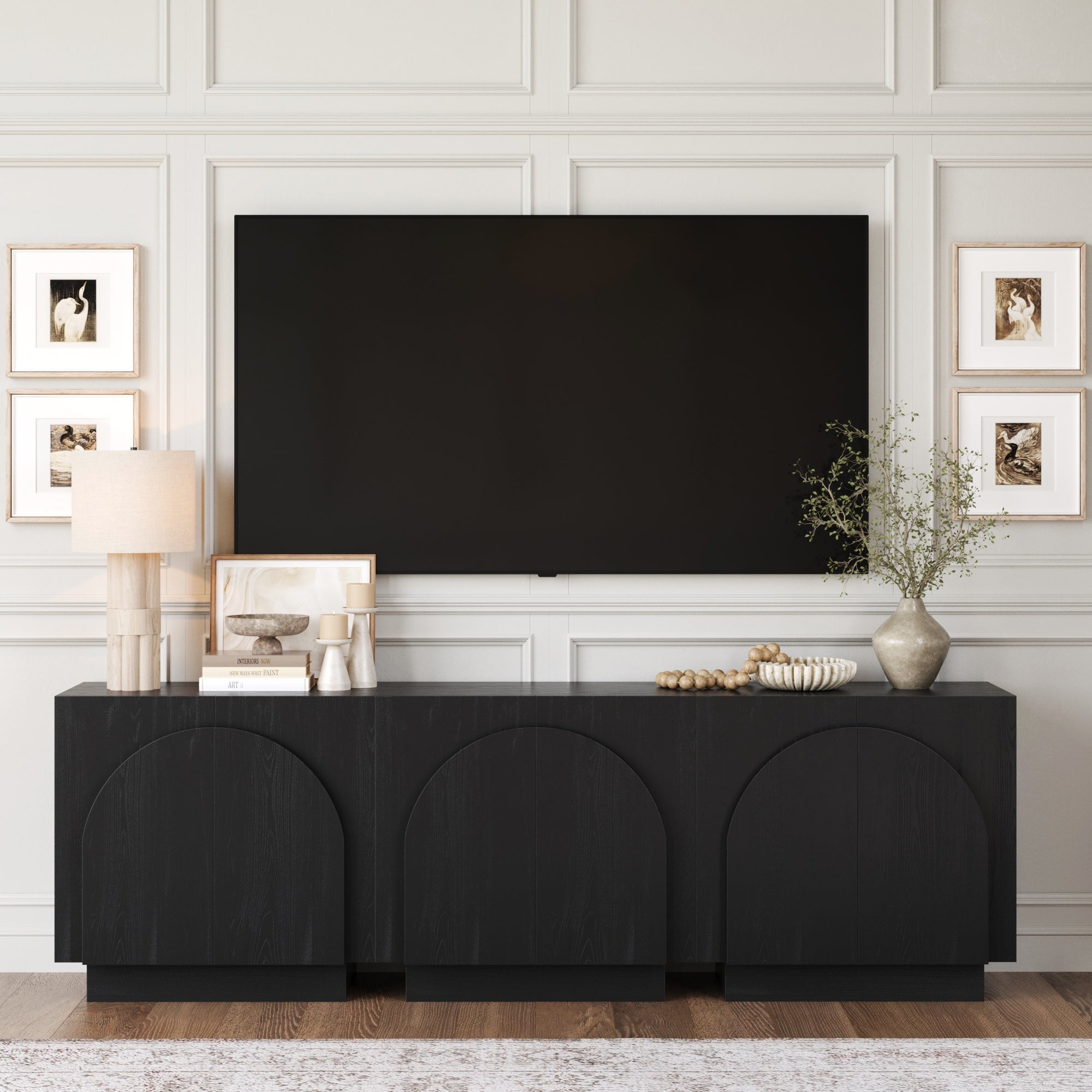 A modern living room features a large TV on a paneled wall above the Nathan James Arched TV Console Credenza Black (Set of 3), styled with a lamp, vases, books, greenery, and four framed art pieces surrounding the TV.
