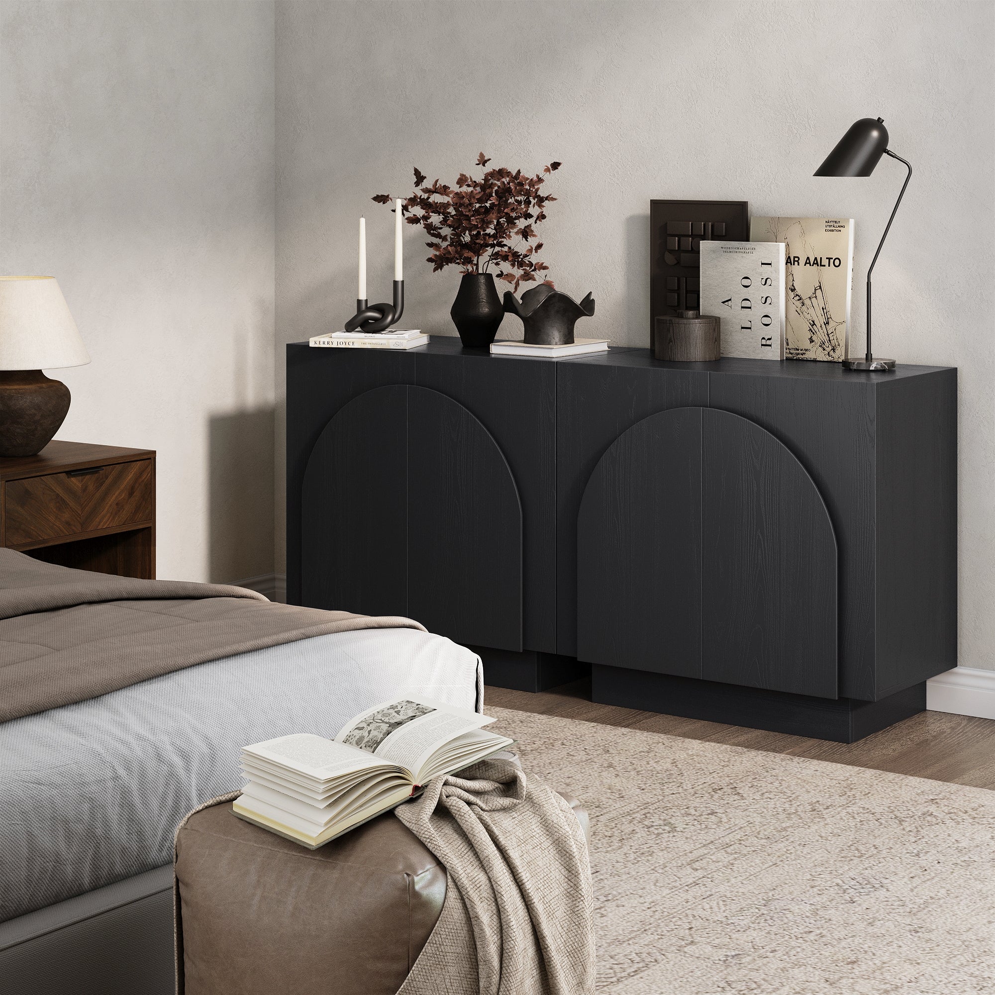 A modern bedroom showcases the Nathan James Wood Mid-Century Arched Storage Cabinet in black, styled with books, a lamp, dried flowers, and decor. A cozy bed and pouf with an open book are set in the inviting foreground.