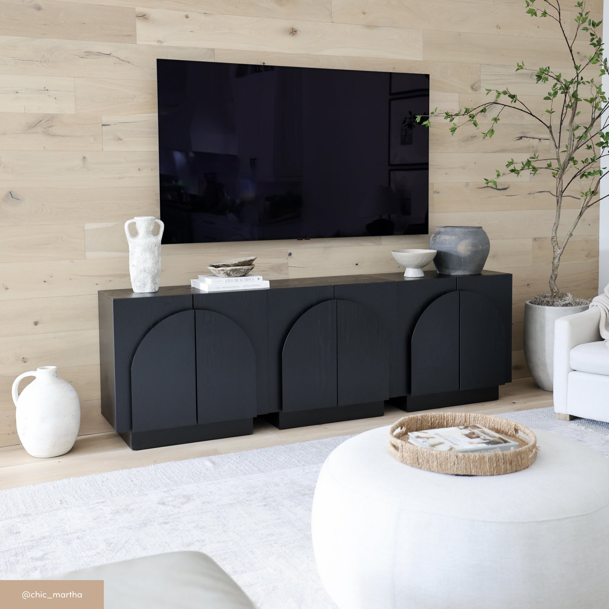 A modern living room features a large wall-mounted TV above a black cabinet with arched details. Minimalist ceramics and a potted plant decorate the space, complemented by light wood-paneled walls and neutral furniture.