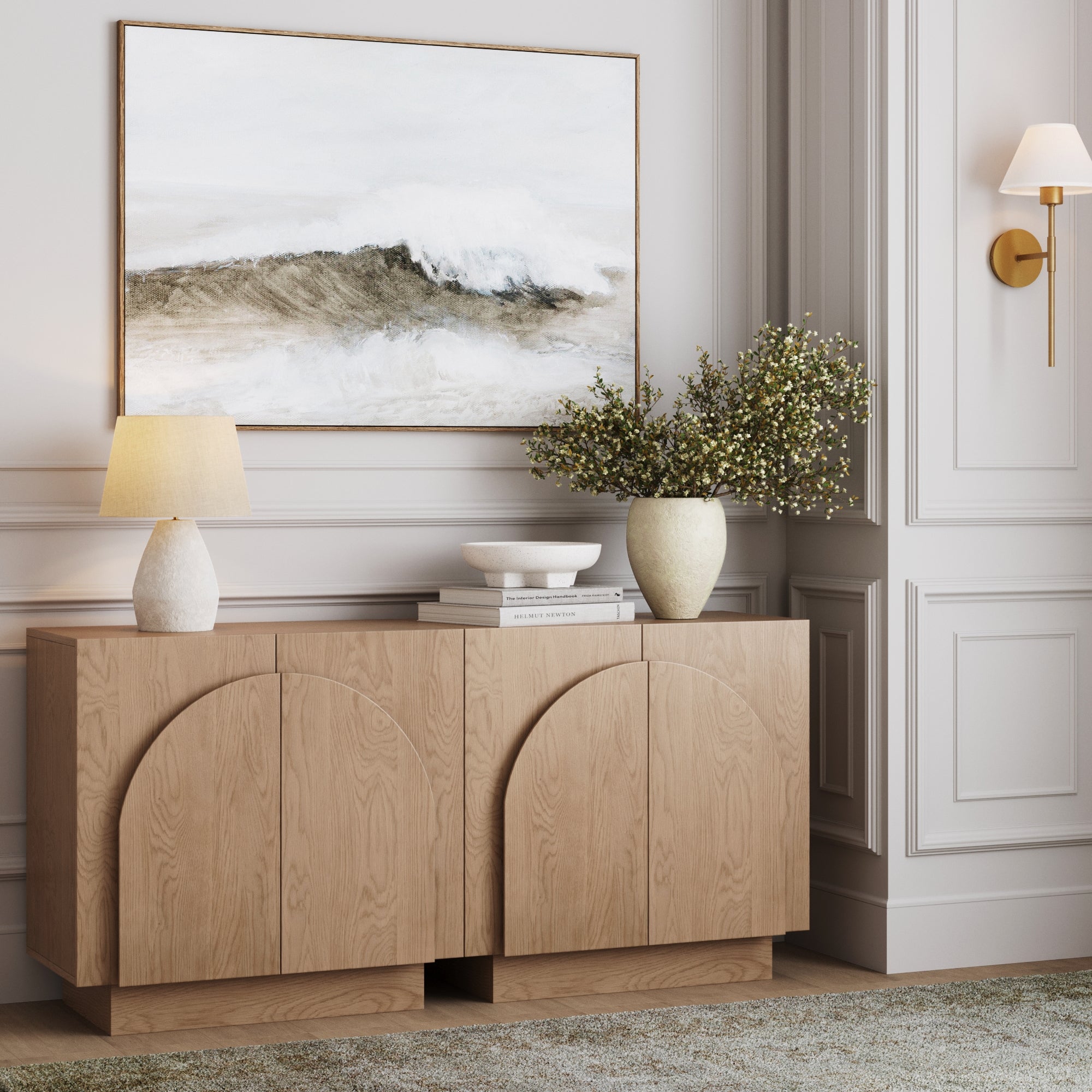A modern wooden sideboard with curved patterns holds a lamp, vase with flowers, and decorative objects. Above it hangs a painting of an ocean wave. The wall features elegant paneling and a brass wall sconce.