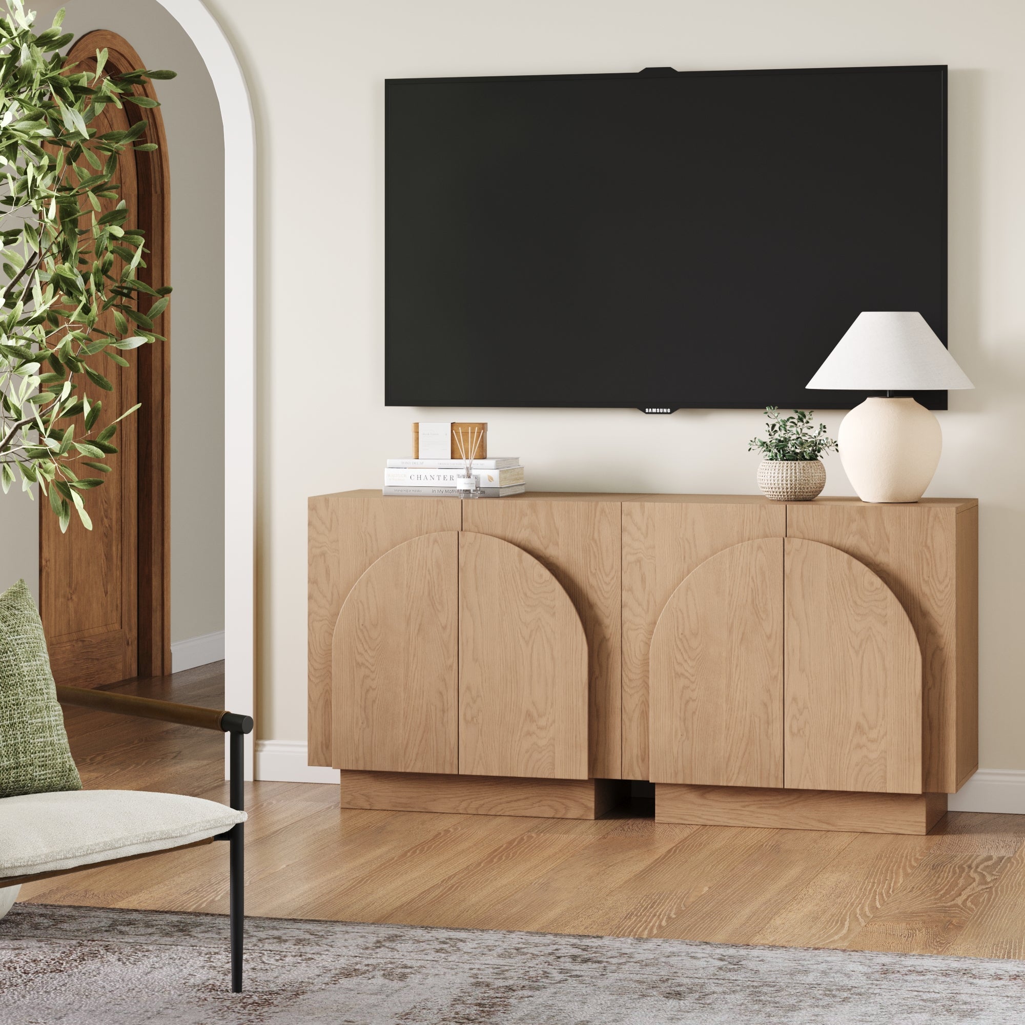 A modern living room showcases the Nathan James Arched TV Console Credenza Oak (Set of 2) beneath a wall-mounted TV, styled with books, a lamp, and a potted plant; a chair and leafy plant add to the stylish décor.