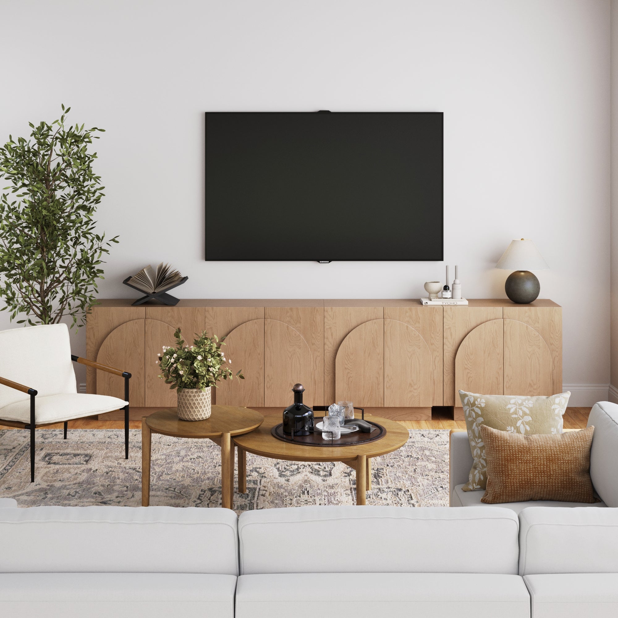 A modern living room showcases a white sofa, patterned rug, wooden coffee tables, potted plant, and natural wood cabinet. The wall-mounted TV sits above the Nathan James Large Arched TV Console Credenza Oak (Set of 4), styled with decor and a lamp.