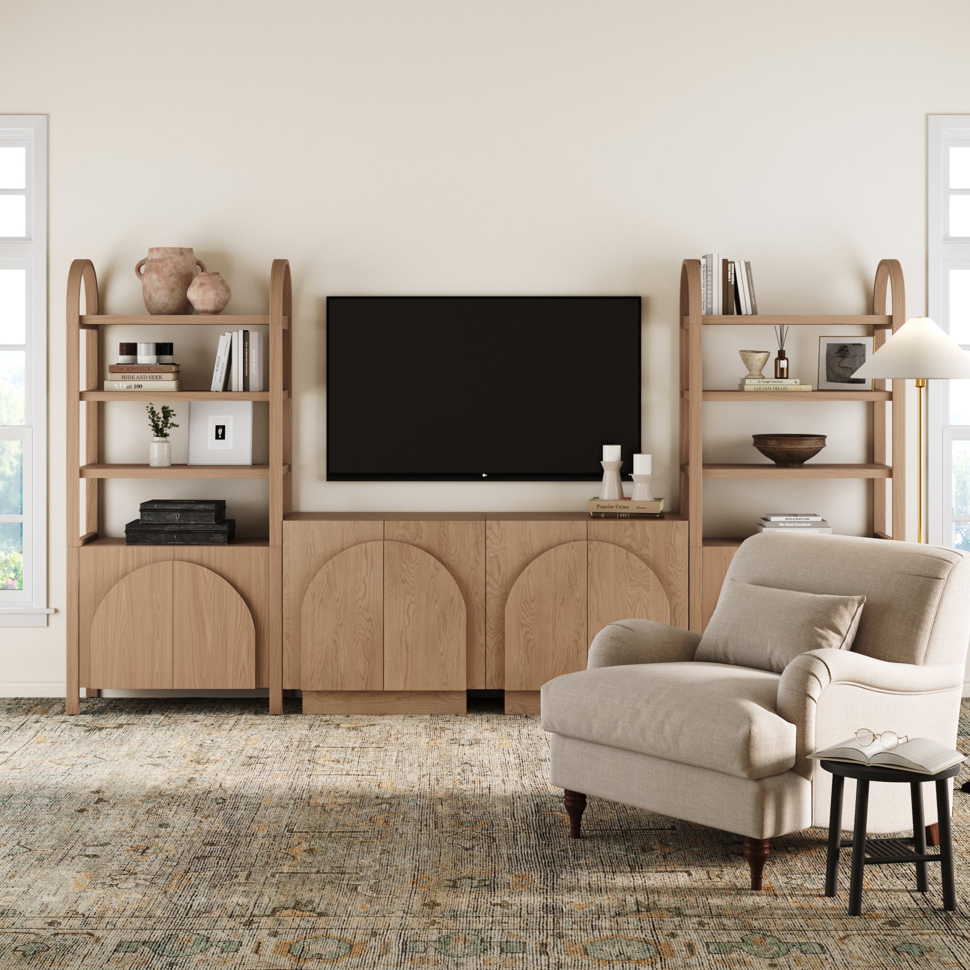 A cozy living room with a beige armchair, round side table, and the Nathan James Mid-Century Modern Arched Wood Entertainment Center (2 Cabinets) displaying decor and a TV, all set on a patterned rug.