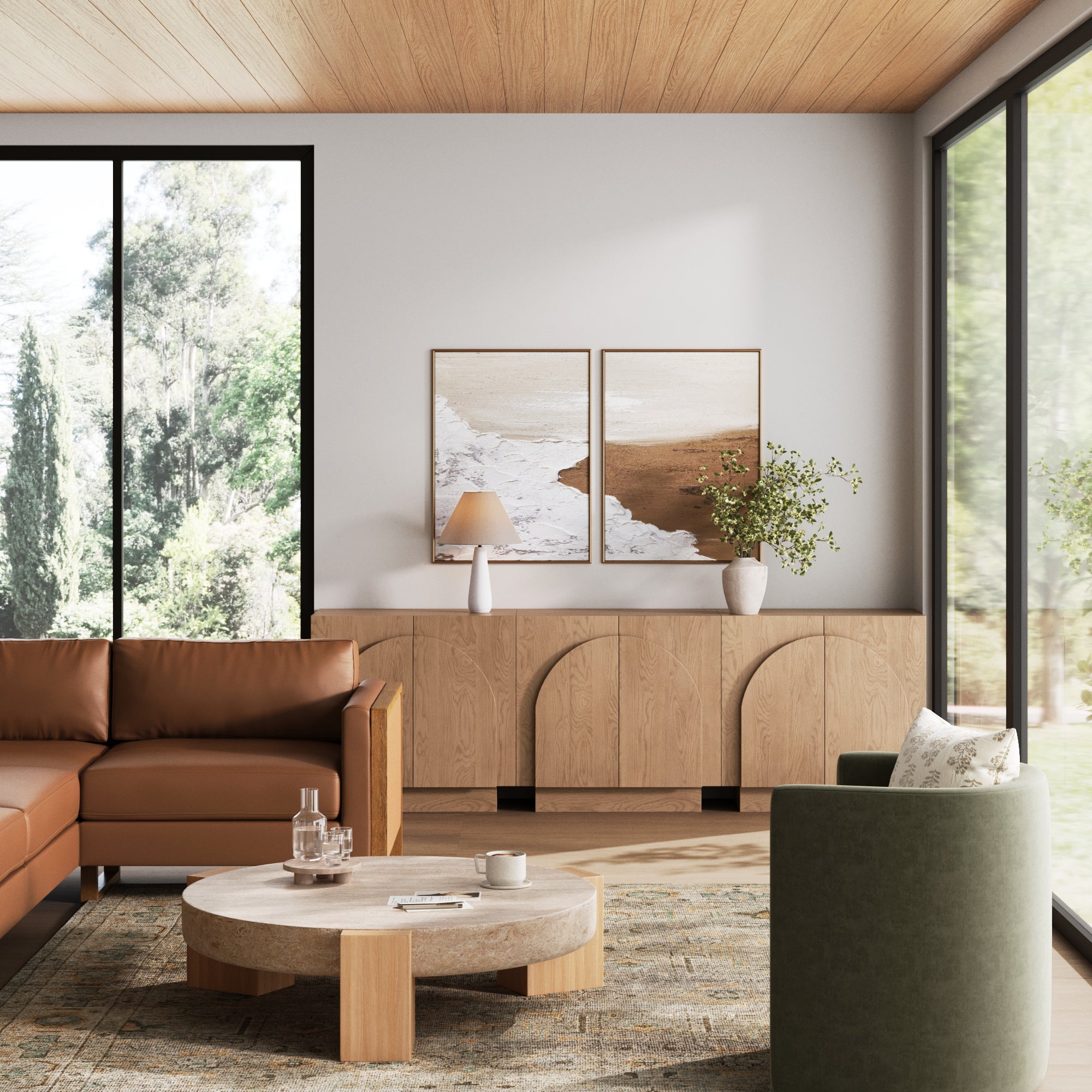 A modern living room with a brown leather sectional, round wooden coffee table, green armchair, large windows, wood ceiling, and a Nathan James Arched TV Console Credenza Oak (Set of 3) topped with beach prints and vases of greenery.