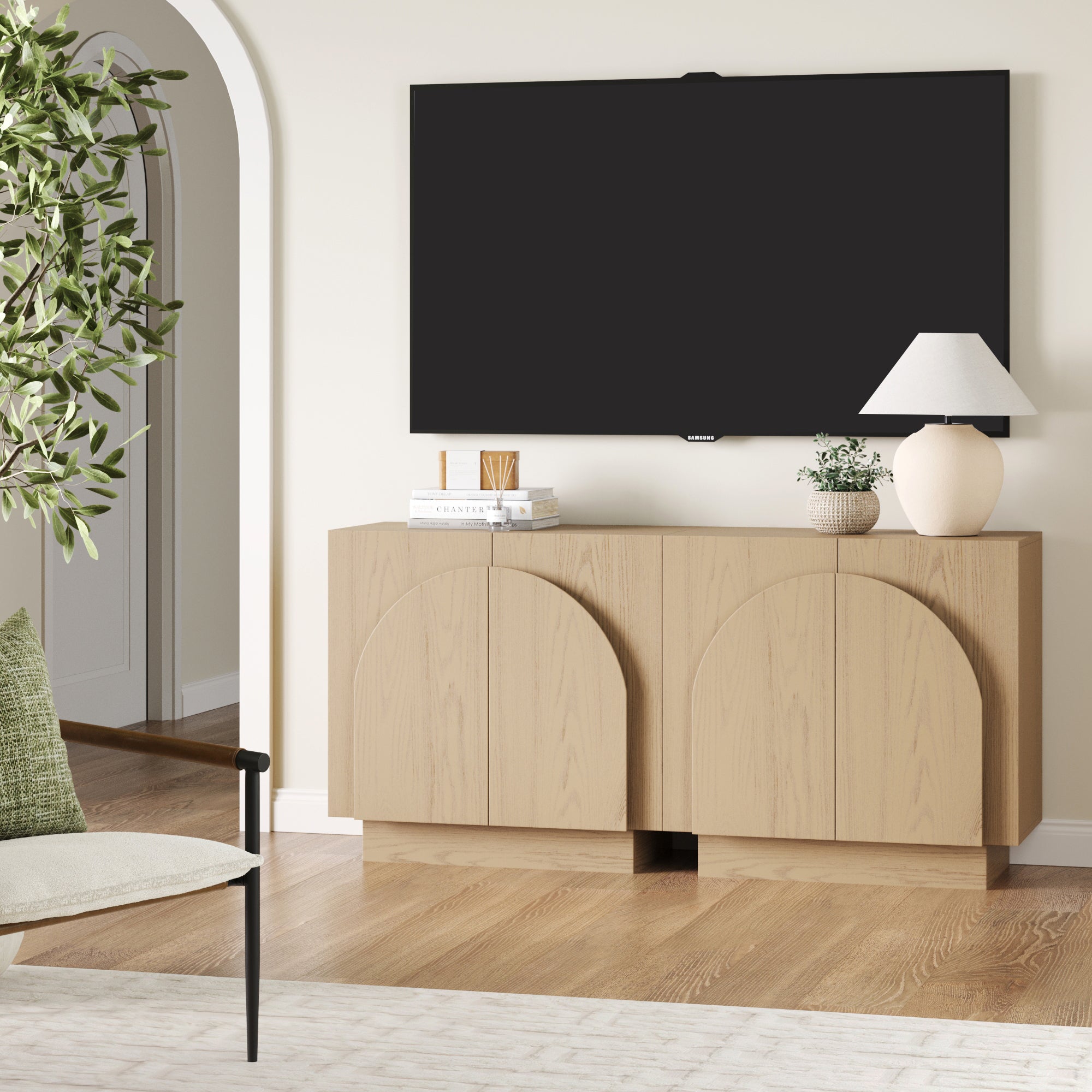 A modern living room features the Nathan James Wood Mid-Century Arched Storage Cabinet in Gray Oak (Set of 2), a white table lamp, books, and a potted plant. A flat-screen TV hangs above, with a green armchair and leafy plant on the left.