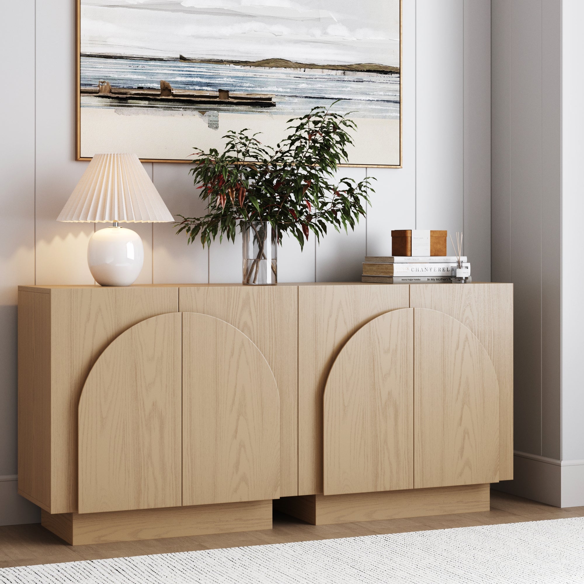 A pair of Nathan James Wood Mid-Century Arched Storage Cabinets in Gray Oak display a white lamp, potted plant, books, and candles. Above hangs a framed coastal painting, with light walls and carpet creating a serene atmosphere.