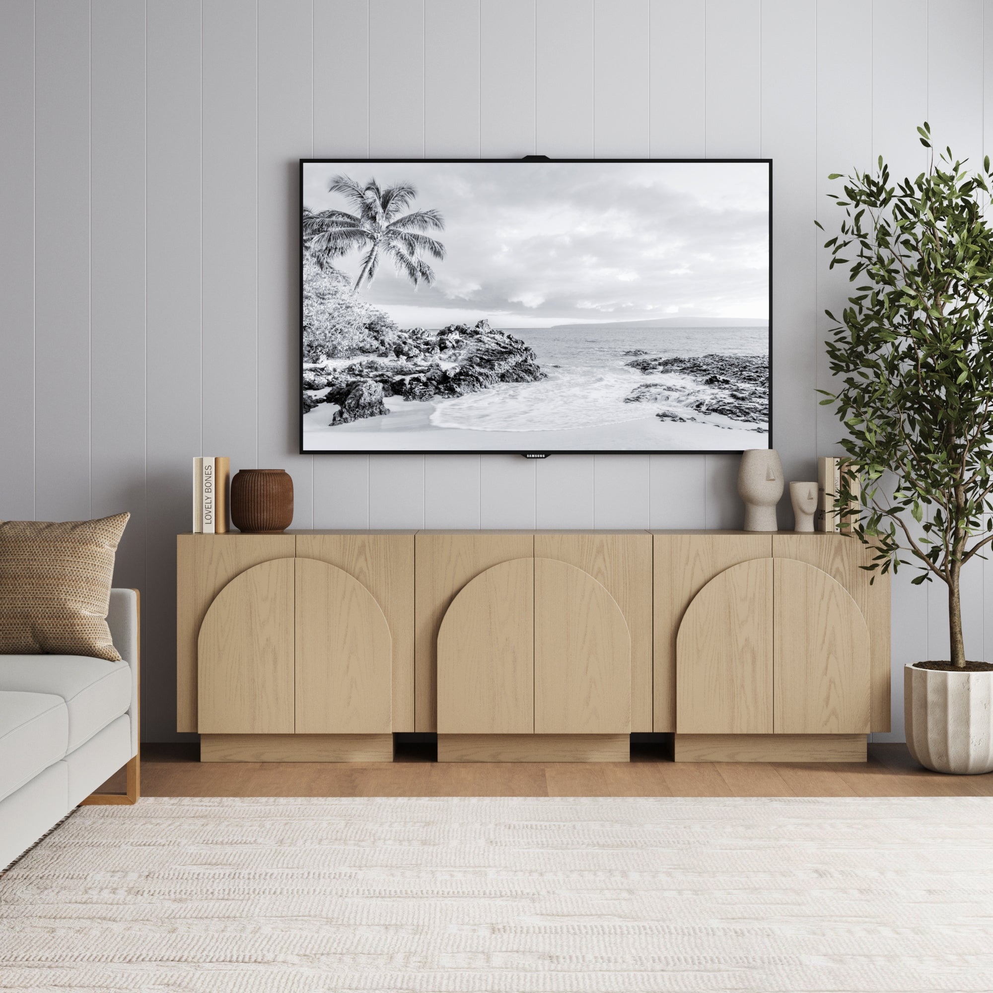 A modern living room with a Nathan James Wood Mid-Century Arched Storage Cabinet in Gray Oak (Set of 3), arched doors, a large framed TV, beige sofa with cushions, potted plant, books, and a light area rug on wooden floors.