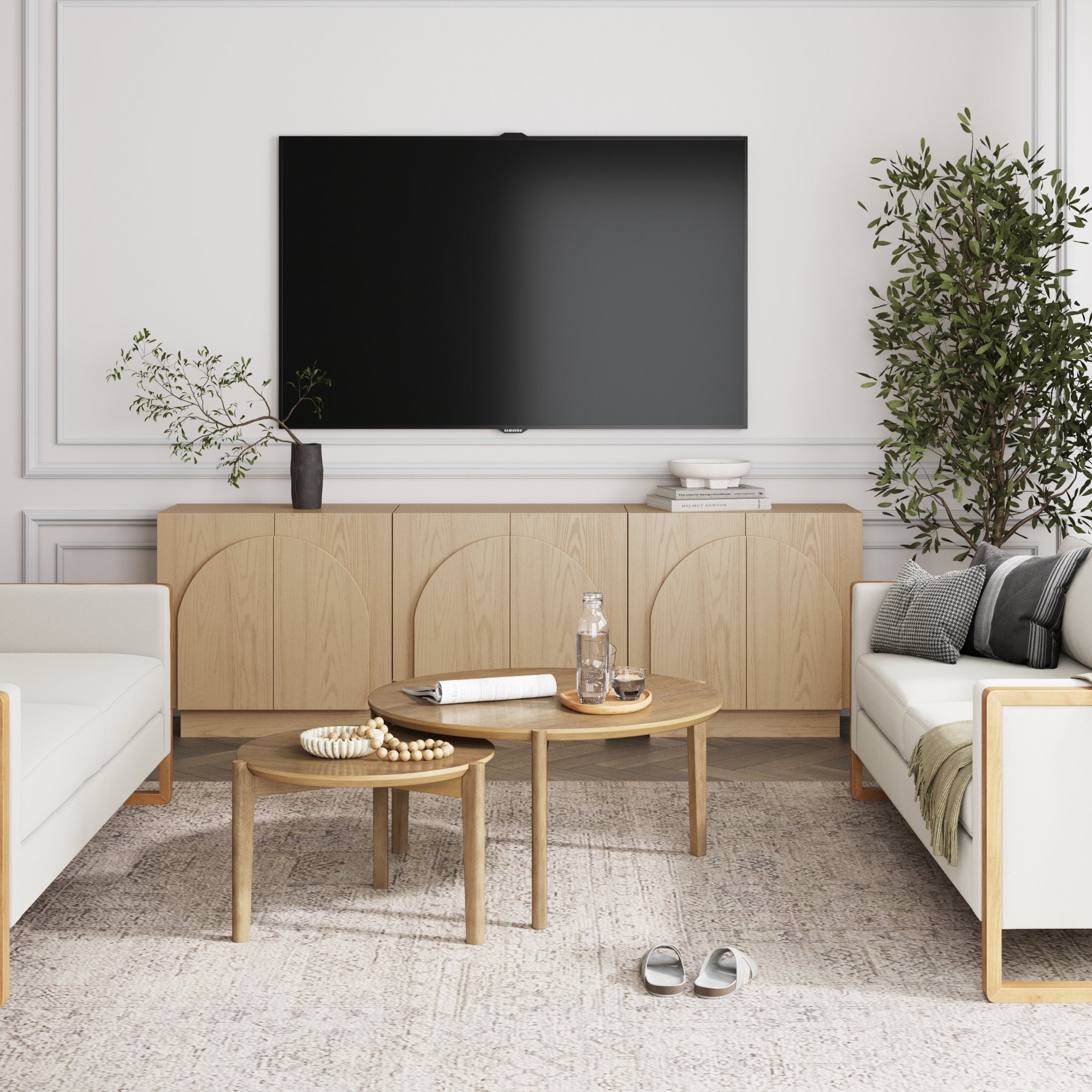 Modern living room with light wood furniture, flat-screen TV, white sofas, round nested coffee tables with glassware, a rug, potted plants, and neutral decor featuring Nathan James Wood Mid-Century Arched Storage Cabinet Gray Oak (Set of 3).