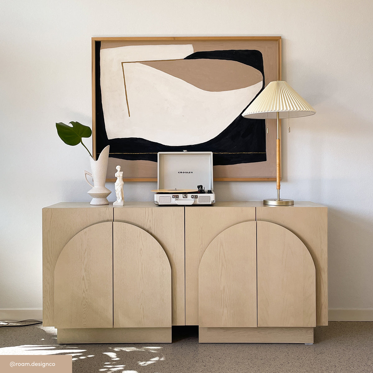 A light wood cabinet with arched doors holds a vase with a leafy branch, a small white statue, a white record player, and a pleated lamp. A modern abstract painting in neutral tones hangs above.