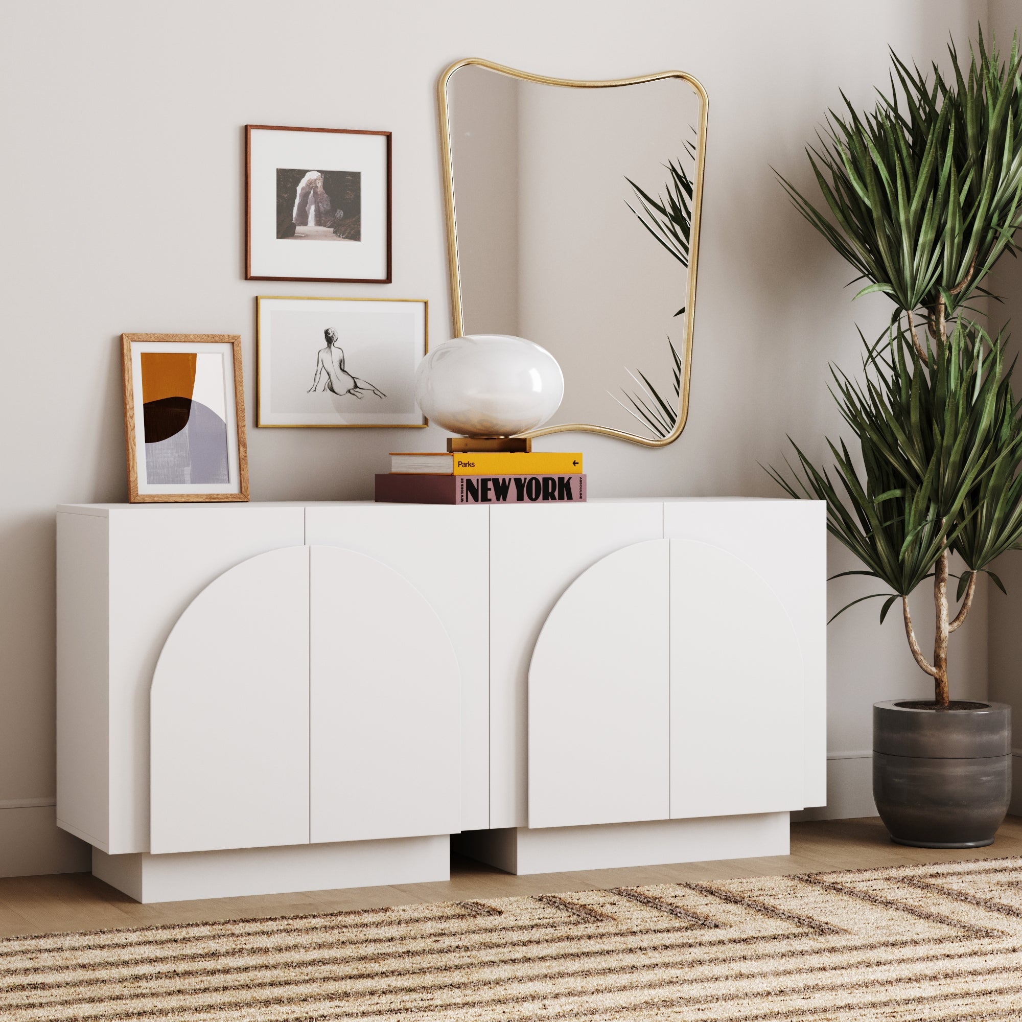 A modern white Nathan James Wood Mid-Century Arched TV Console Credenza with two cabinets stands on a striped rug, topped with framed art, a lamp, and books. A gold-framed mirror and tall potted plant accent the light, minimalist space.