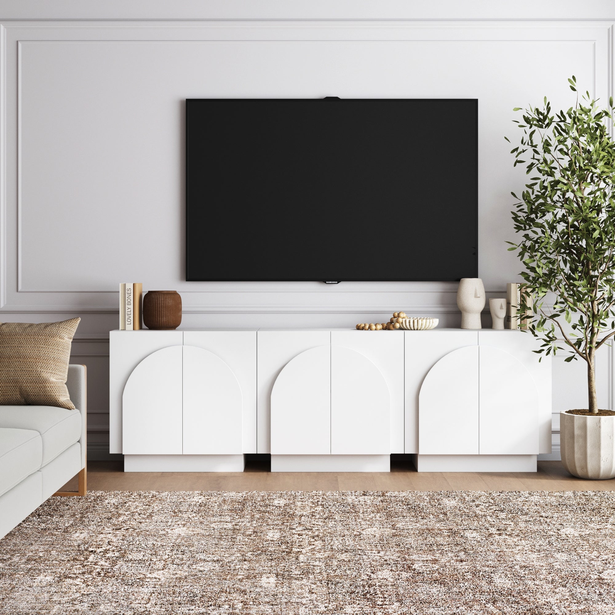 A modern living room has a wall-mounted TV above the Nathan James Wood Mid-Century Arched TV Console Credenza in white with 3 cabinets, styled with books and decor. A beige sofa, textured rug, and tall potted plant create an inviting atmosphere.