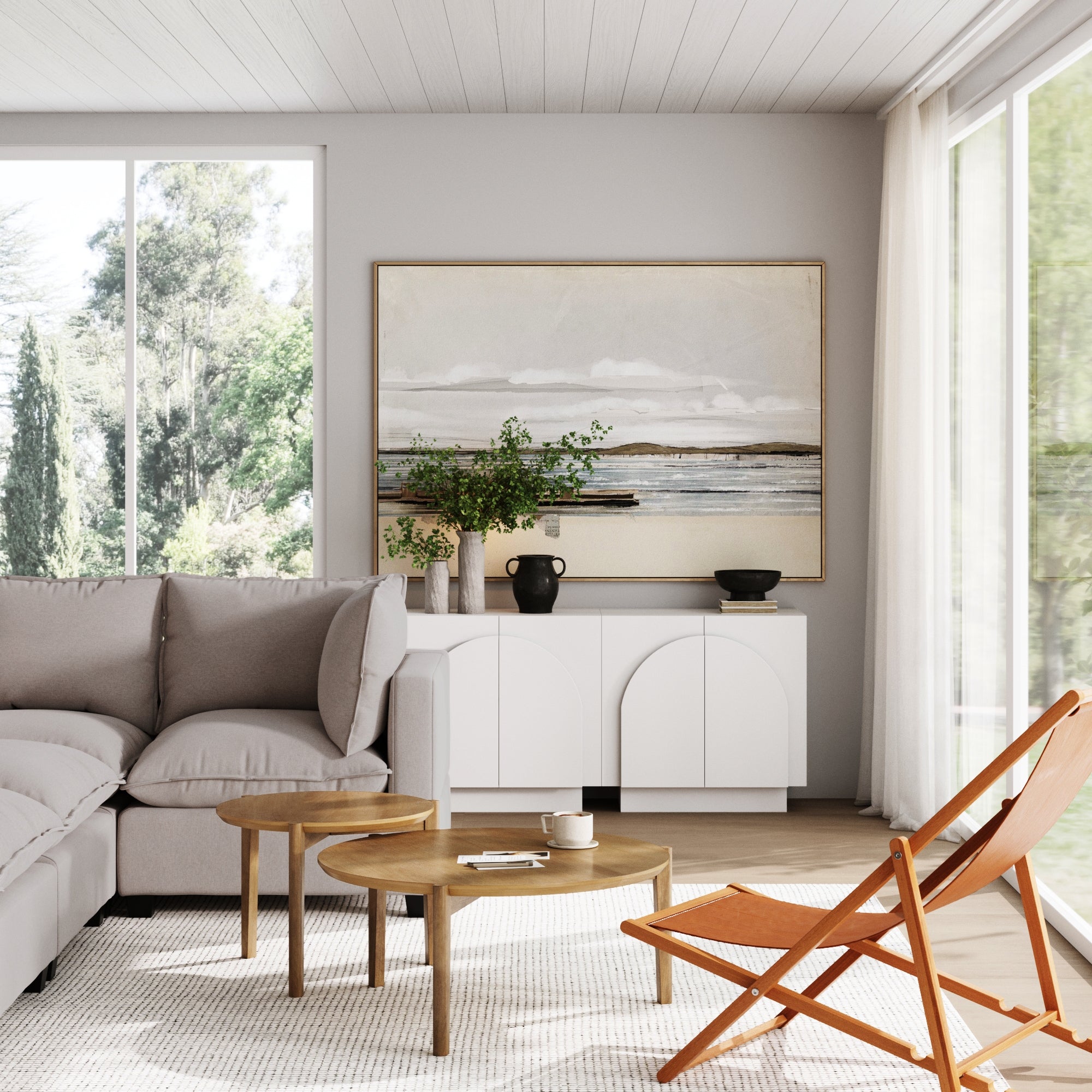 A bright, modern living room with a gray sectional, wooden coffee tables, tan chair, green plants, large windows, and a Nathan James Wood Mid-Century Arched Storage Cabinet White featuring arched doors and an adjustable shelf.