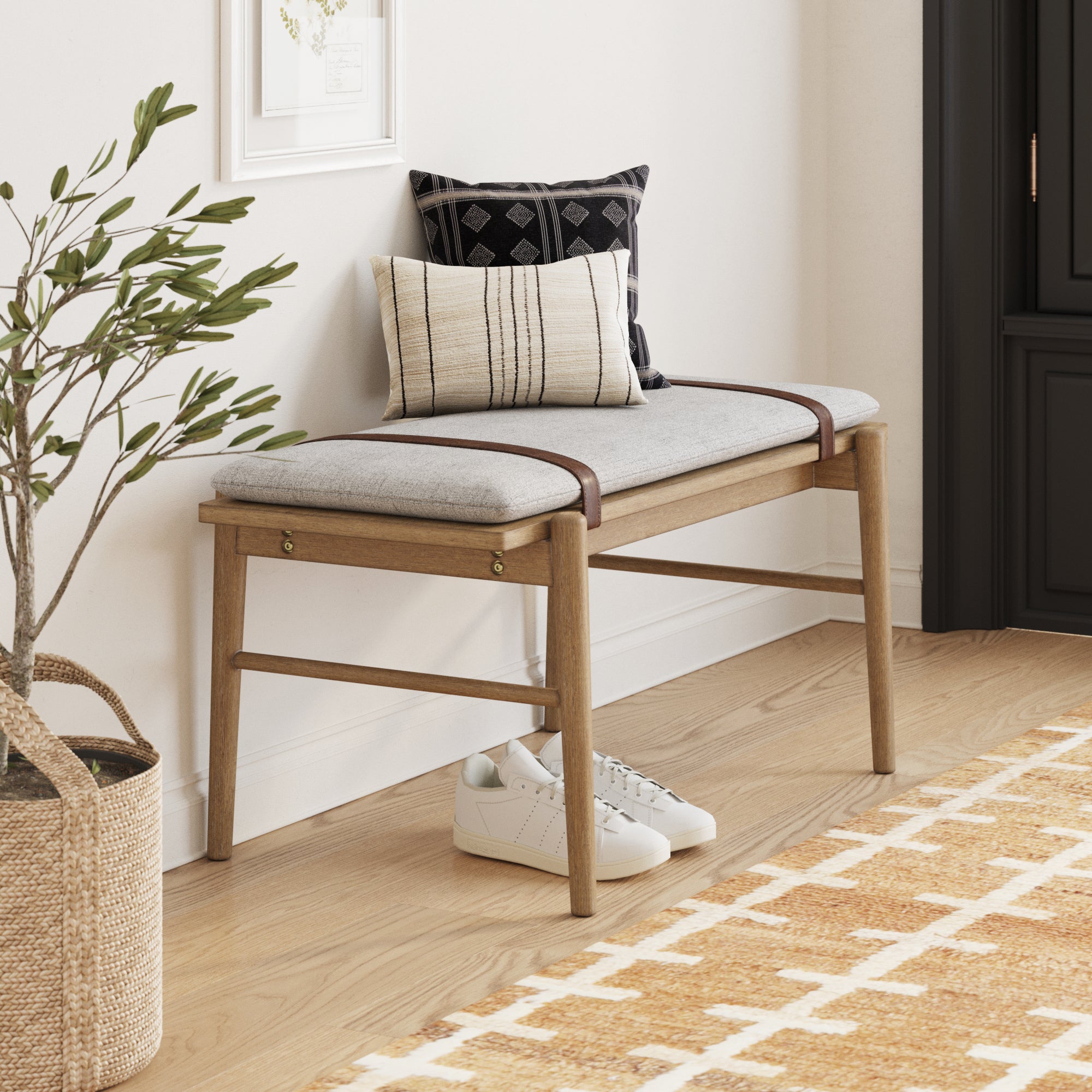 A modern wooden bench with a grey cushion, two decorative pillows, and leather straps sits against a white wall. White sneakers are placed underneath, and a woven basket with a leafy plant is nearby on a patterned rug.