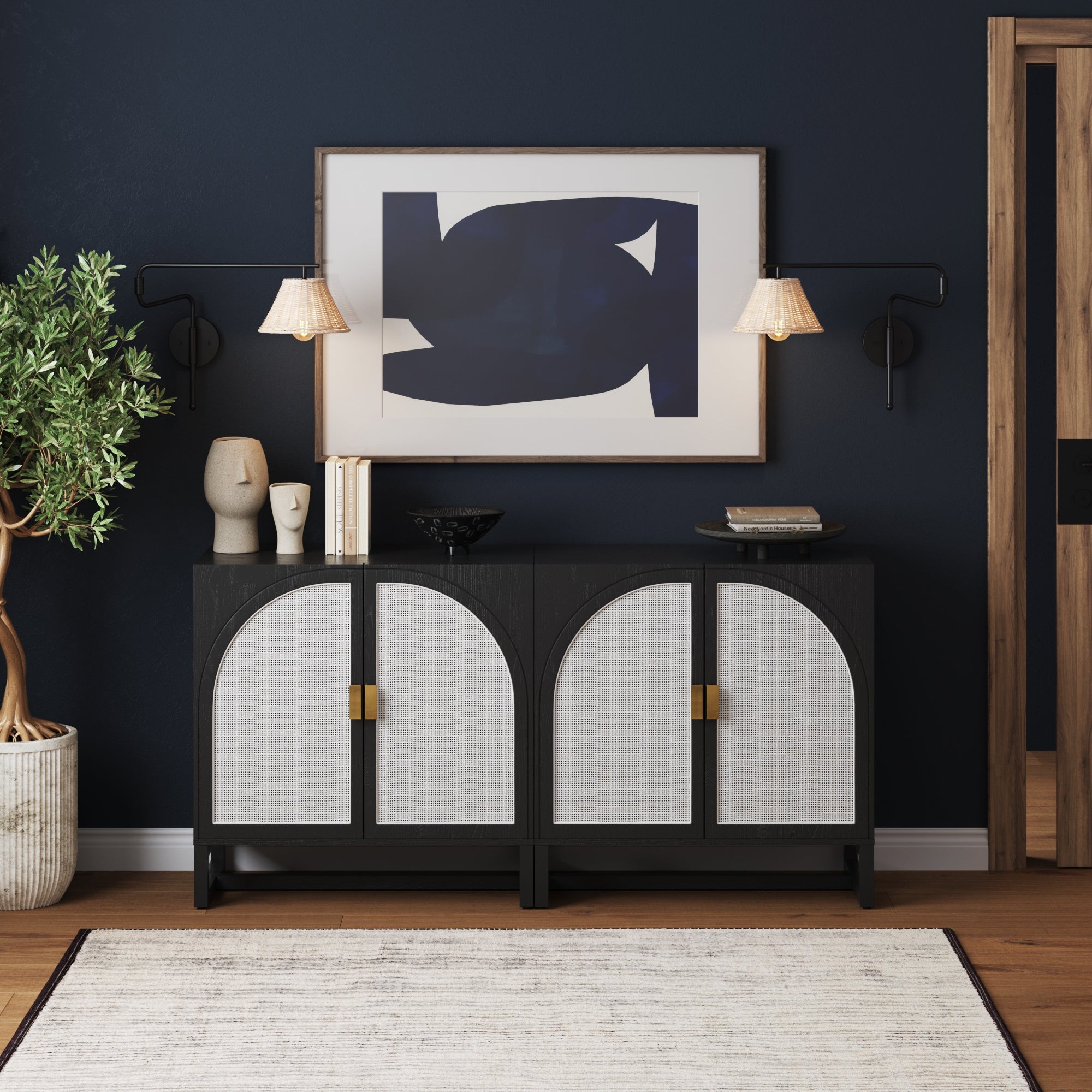 A modern console table with arched doors stands against a dark blue wall, topped with books, decorative objects, and a black bowl. Above it hangs abstract art and two wall-mounted lamps. A potted plant is on the left.
