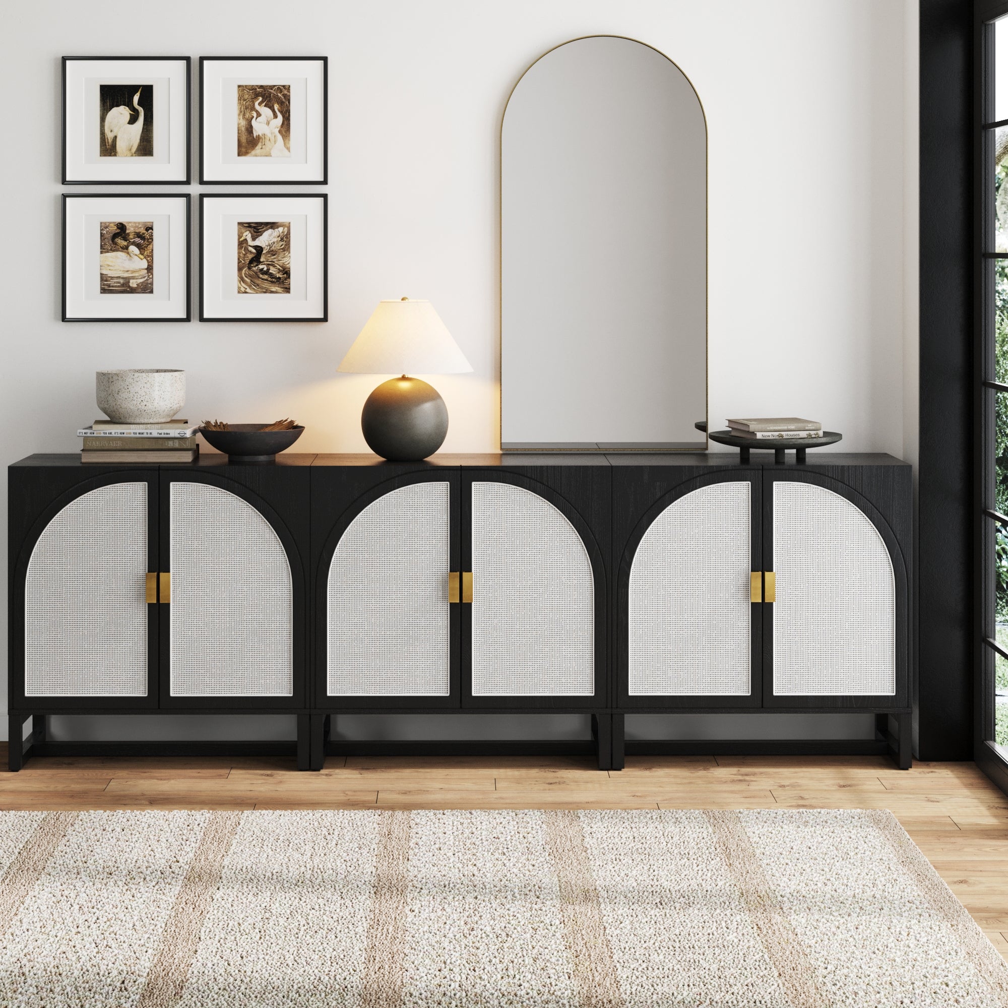 The Nathan James Rattan Arched TV Console Credenza Black (Set of 3) sits against a white wall, featuring arched doors and topped with a lamp, decor bowls, and a large arched mirror. Four framed artworks hang above, with a neutral rug on the wooden floor.