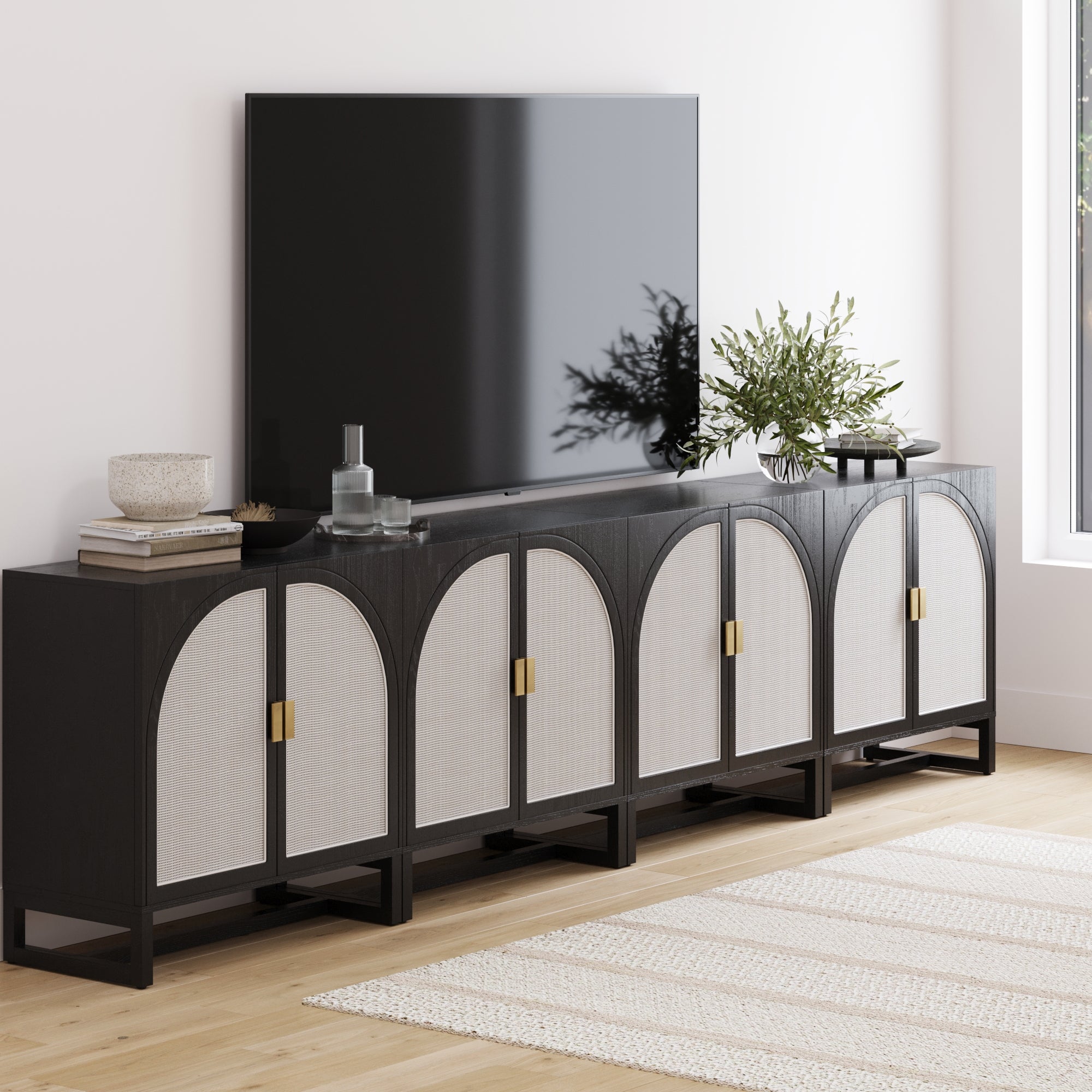 A modern living room featuring the Nathan James Large Rattan Credenza Console in black, set beneath a wall-mounted flat-screen TV and styled with decorative items and greenery on a light wood floor with a cream rug.