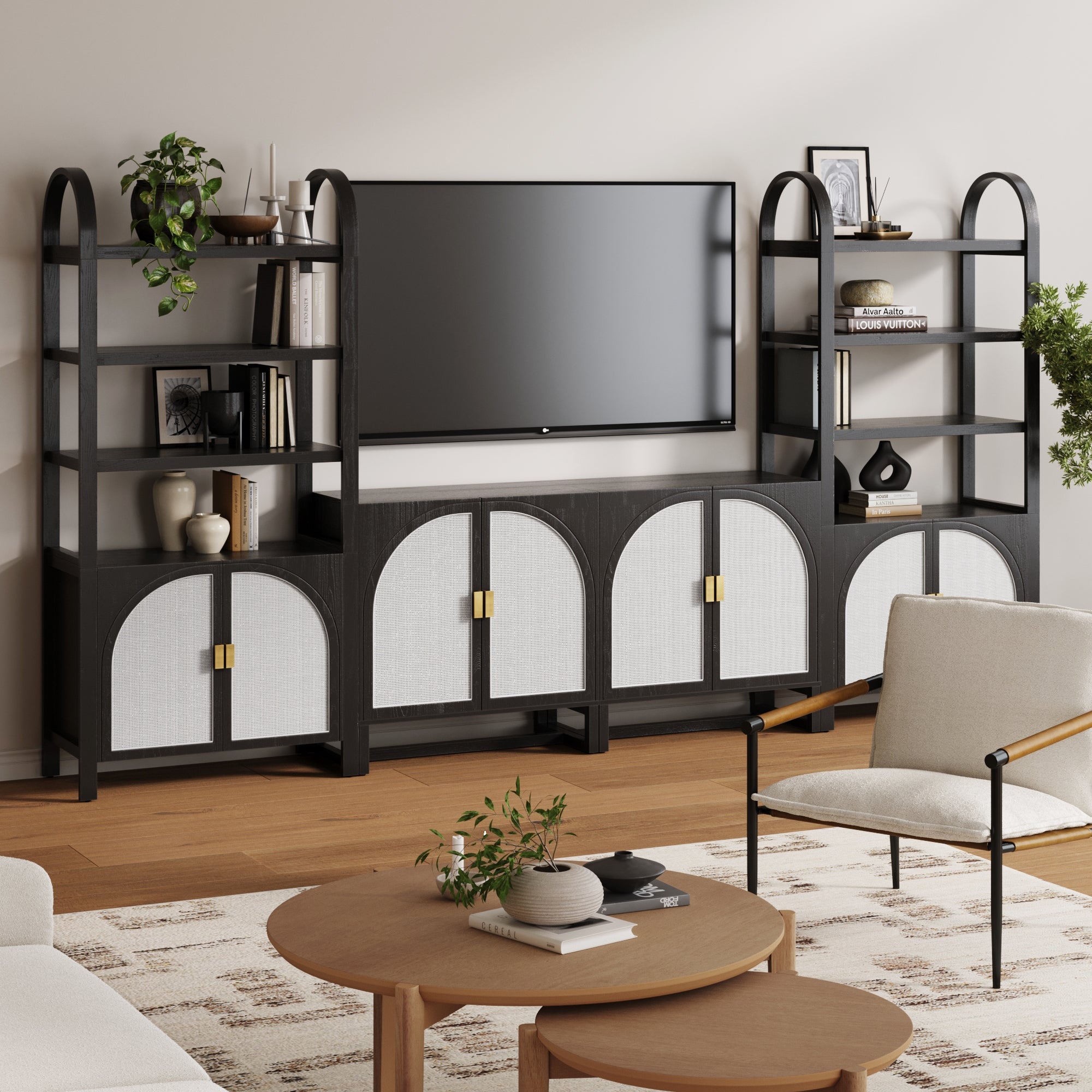 A modern living room with the Nathan James Boho Arched Bookshelf & Cabinet Set TV Stand in black, open shelving for decor, a wall-mounted TV, a round wood coffee table, light armchair, patterned rug on wood floors, and faux rattan storage unit for texture.