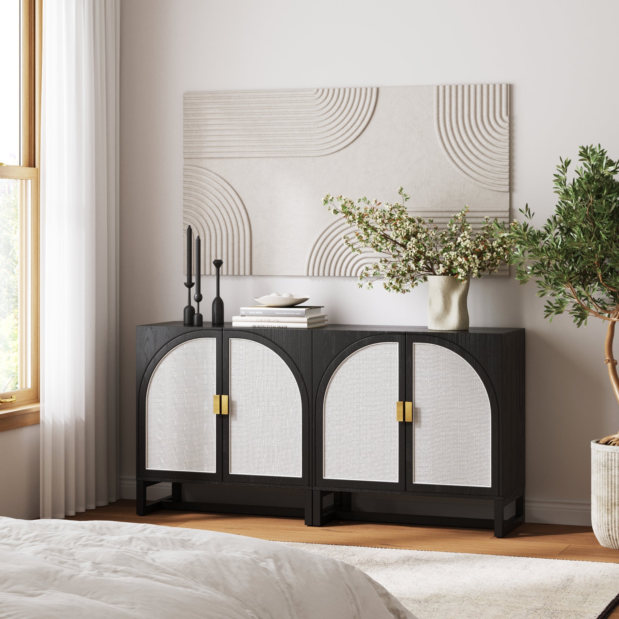 A modern bedroom showcases the Nathan James Boho Rattan Arched Door Storage Cabinet in black, paired with a vase of flowers, two black candlesticks, curved line wall art, a large potted plant, and sunlight streaming through the window.